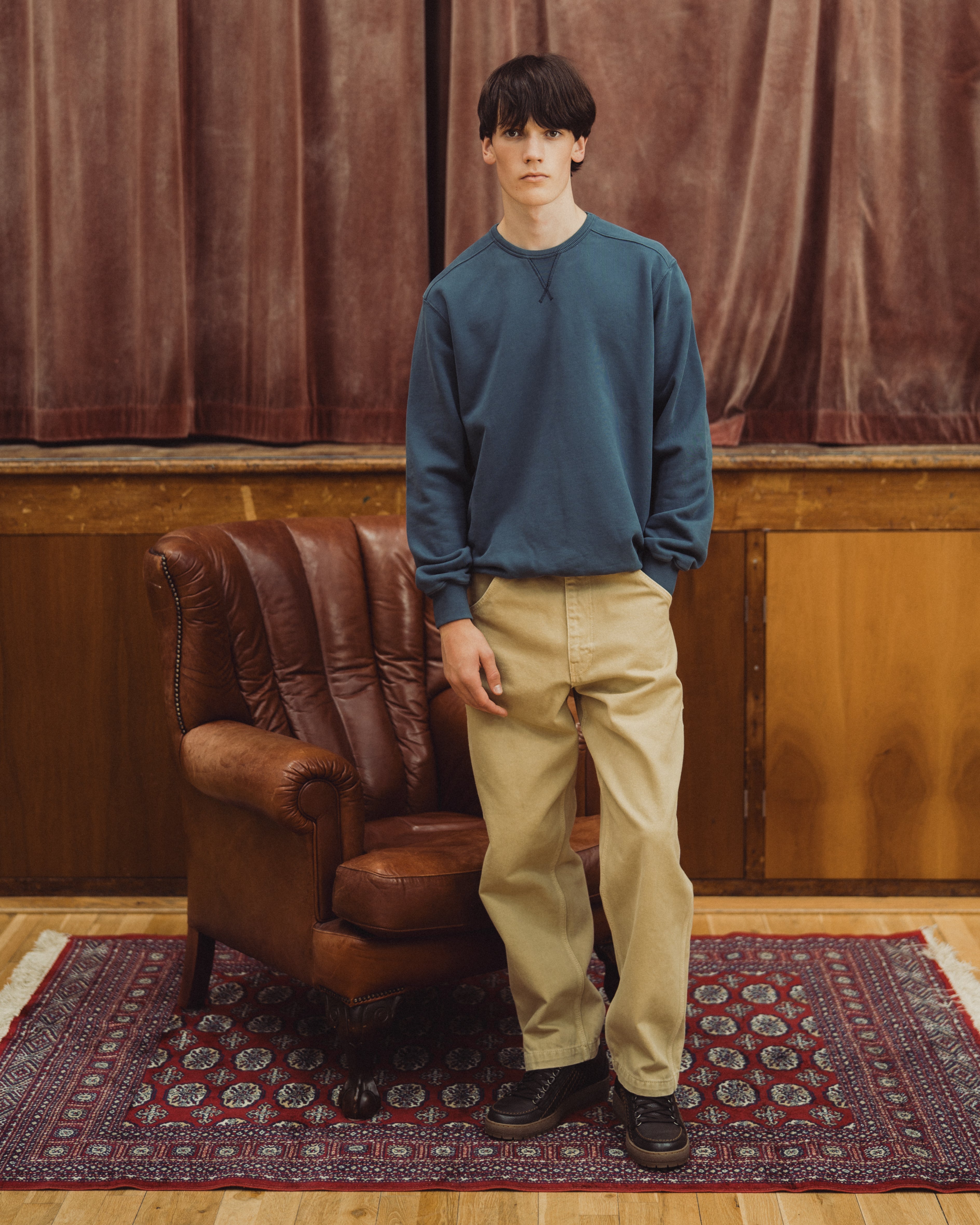 Blue organic cotton sweat , model standing in front of brown leather armchair