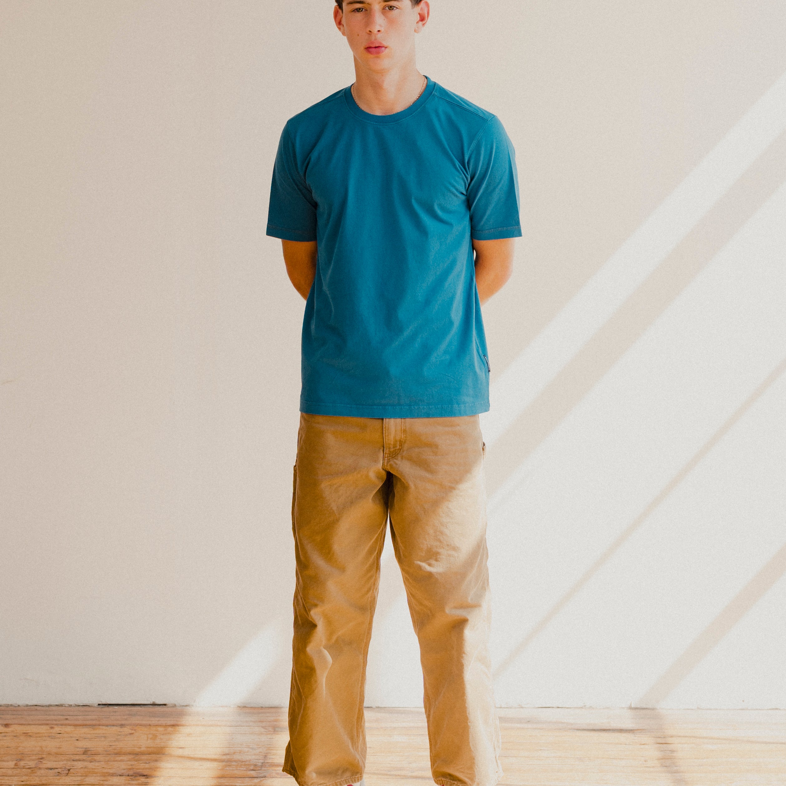 Man wearing a blue t-shirt and beige pants standing in a room with wooden floor and white walls.