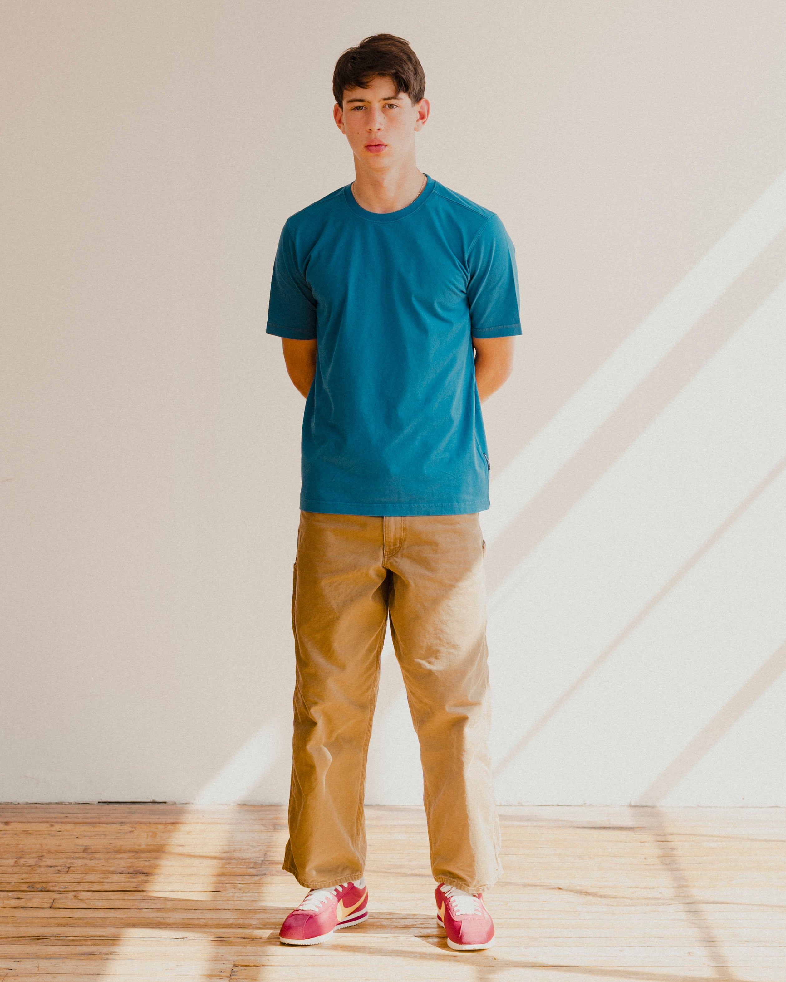 Man wearing a blue t-shirt and beige pants standing in a room with wooden floor and white walls.