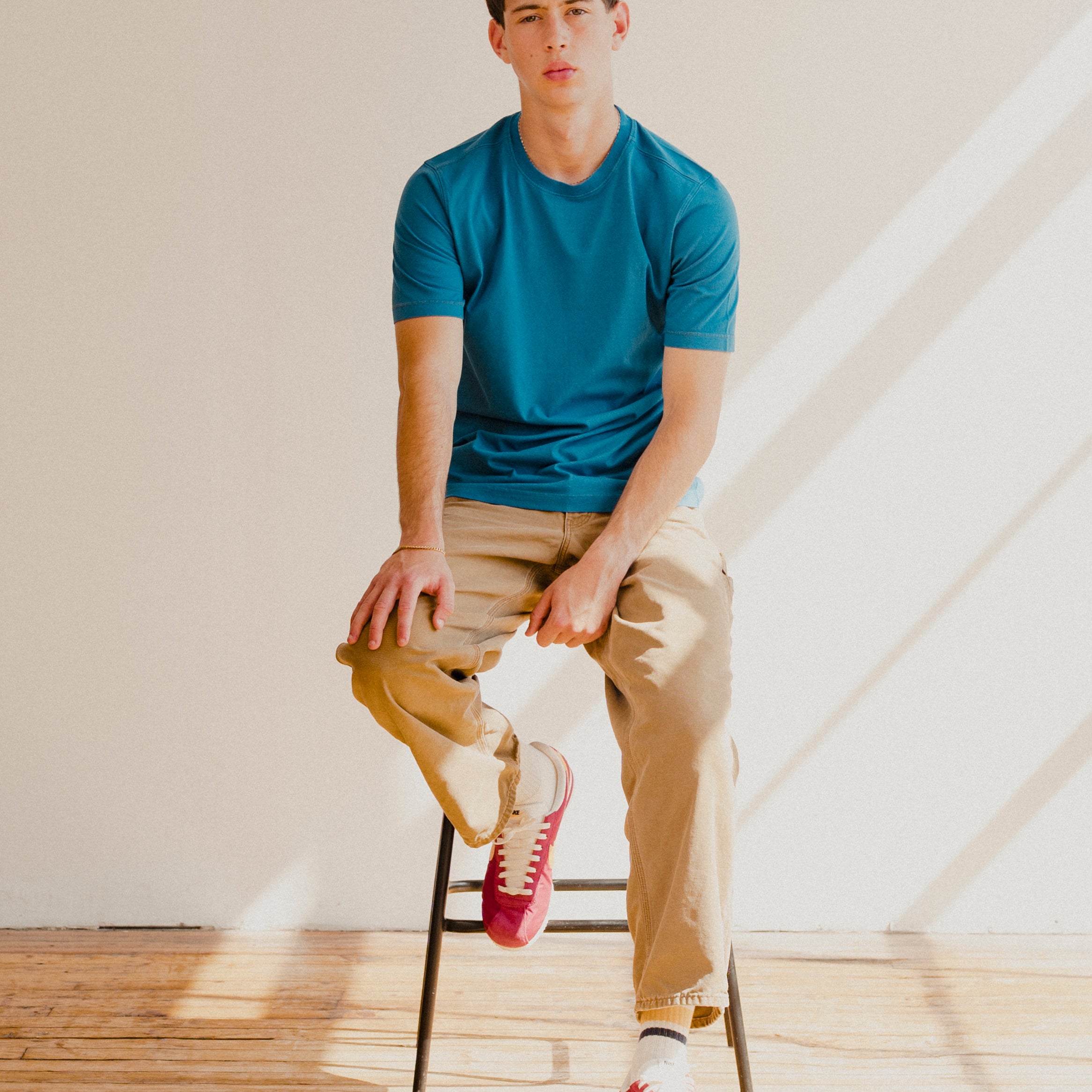 Man sitting on a stool wearing a blue t-shirt, beige pants, and red sneakers in a well-lit room.
