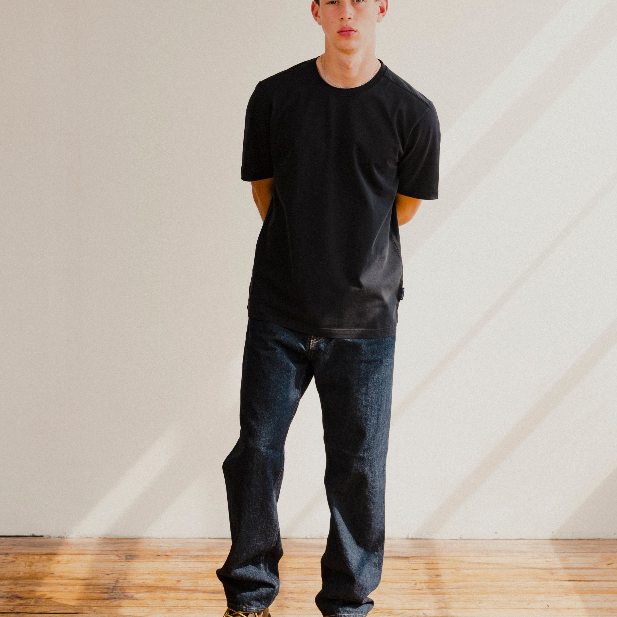 Man wearing a black t-shirt and blue jeans standing against a plain wall.