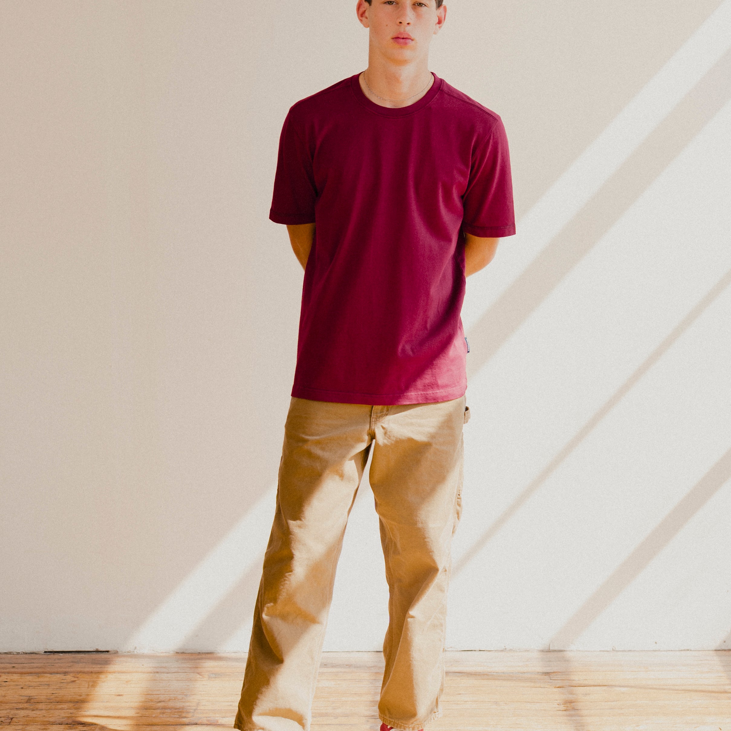 Man wearing a maroon red t-shirt and beige pants standing in a room with sunlight streaming through a window.