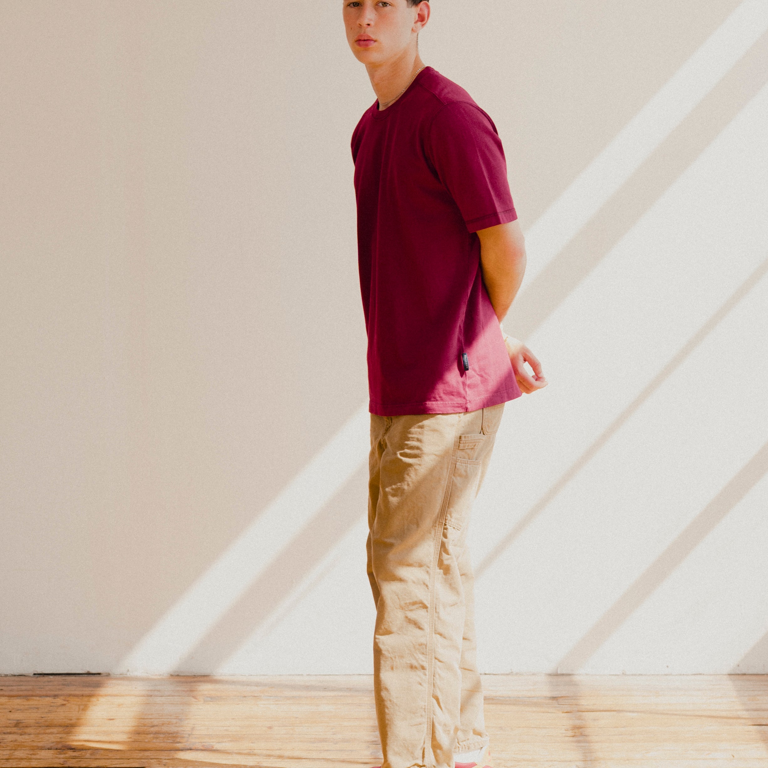 Man wearing a maroon red shirt and beige pants standing in a room with wooden floor and white walls.