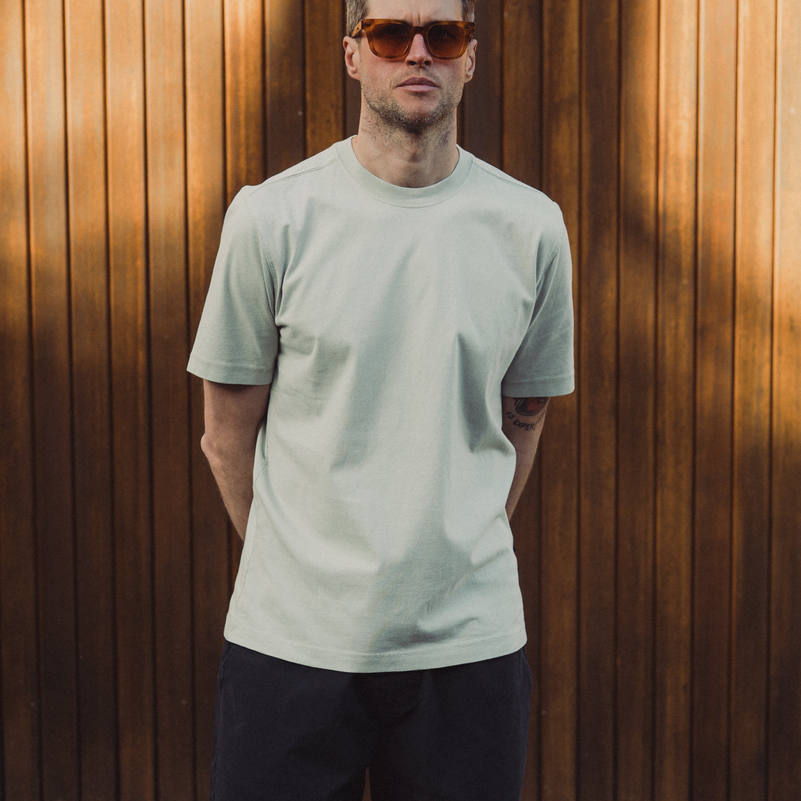 Man wearing a light green t-shirt and sunglasses standing against a wooden wall.