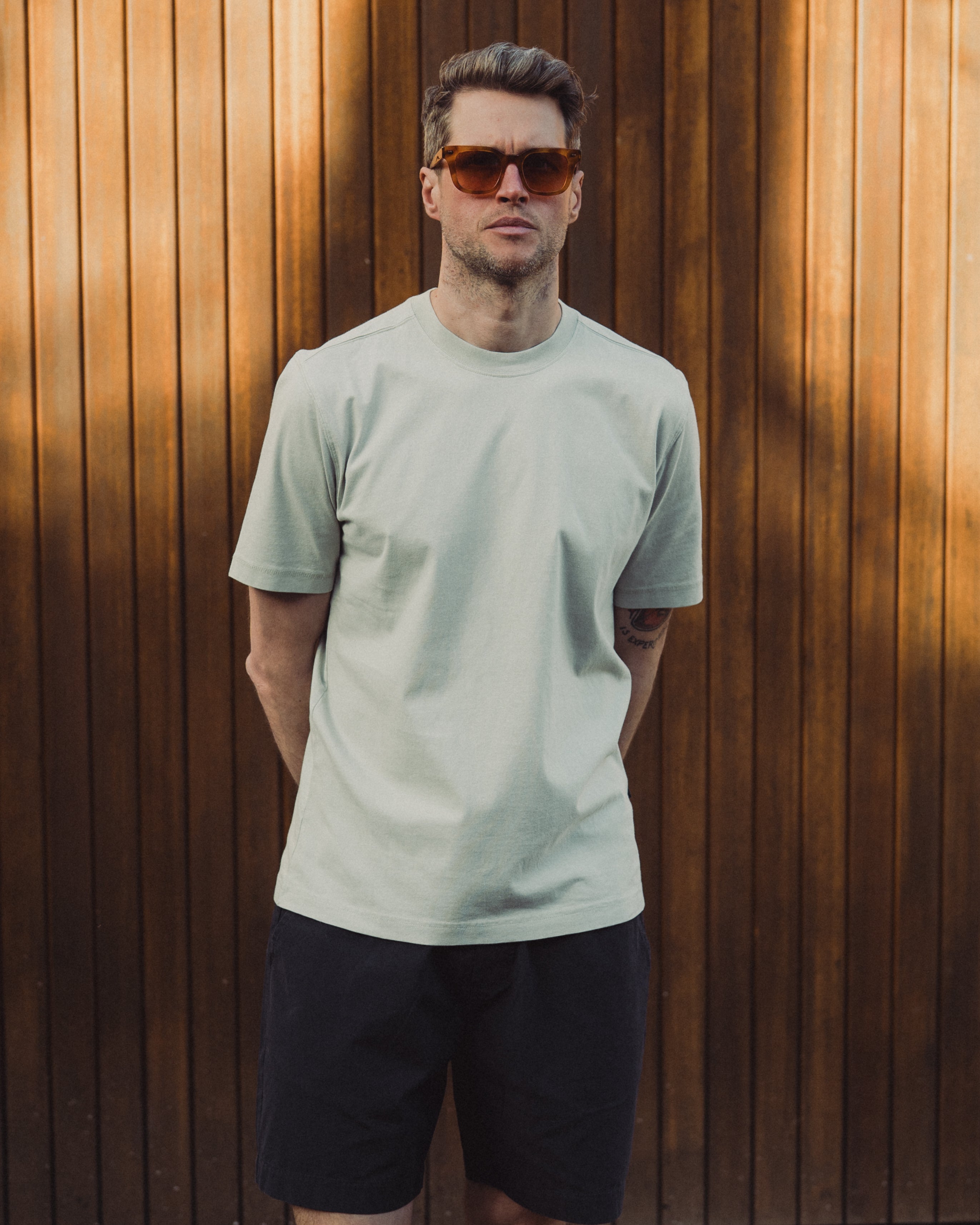Man wearing a light green t-shirt and sunglasses standing against a wooden wall.