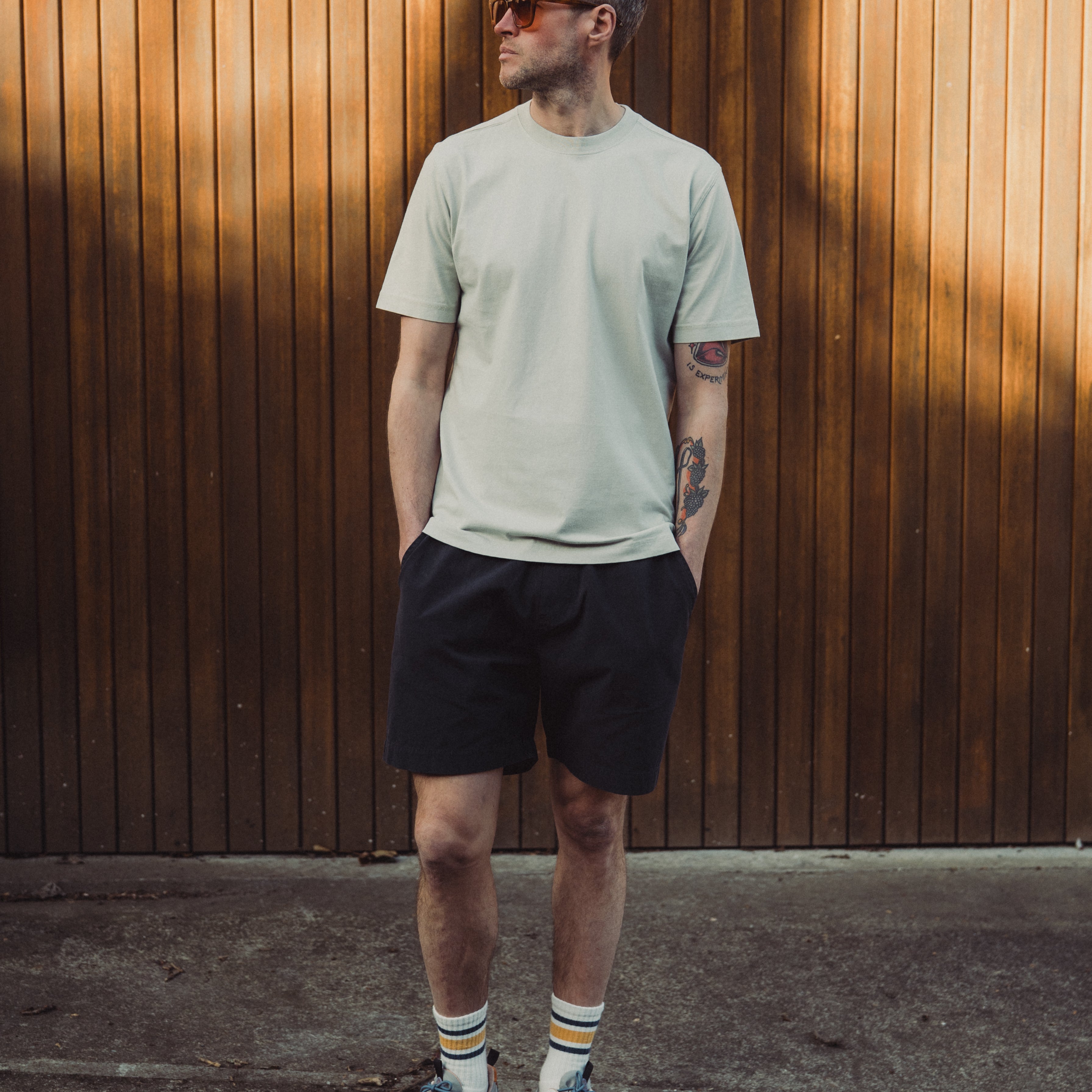 Man wearing a light green t-shirt and black shorts standing against a wooden wall.