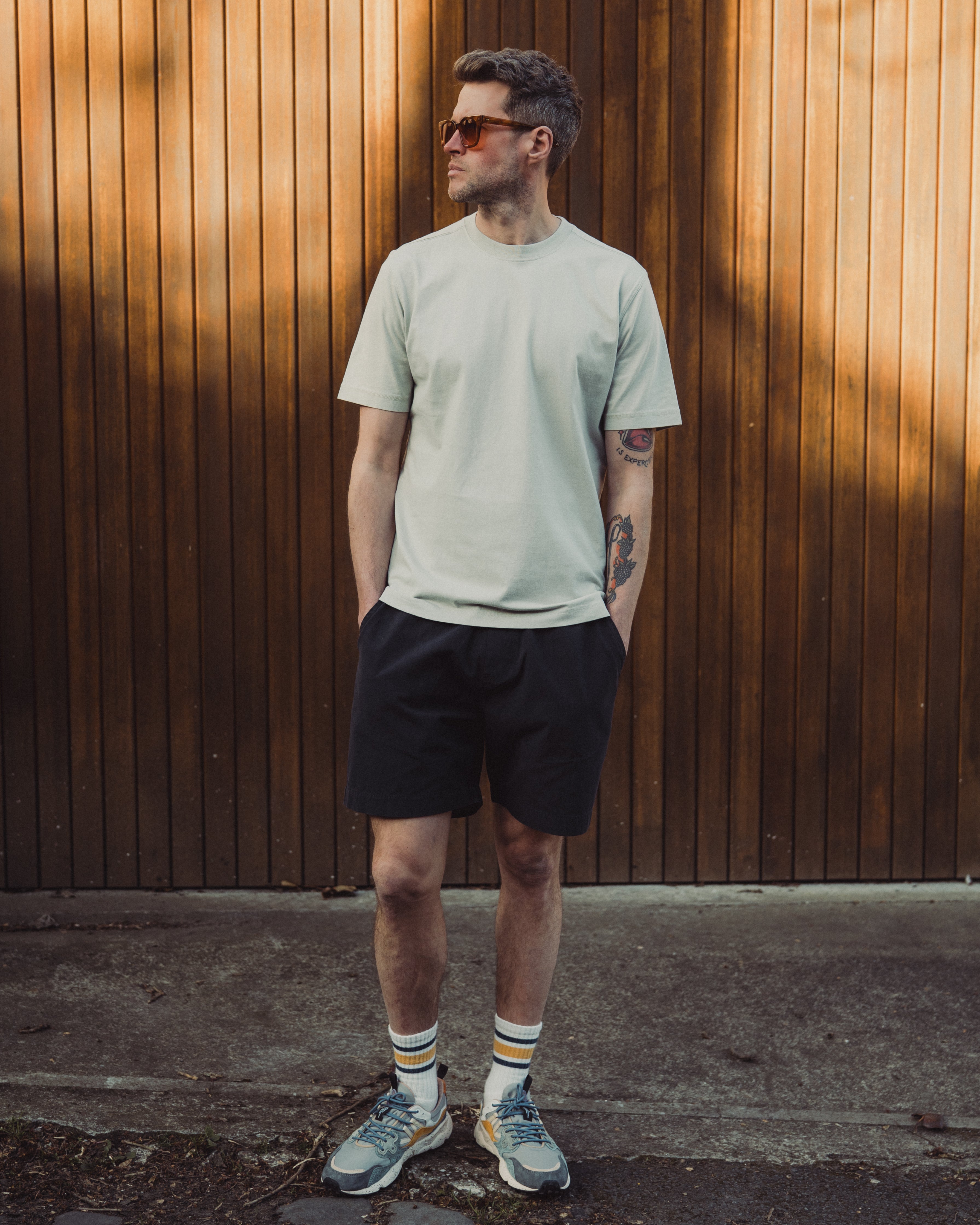 Man wearing a light green t-shirt and black shorts standing against a wooden wall.