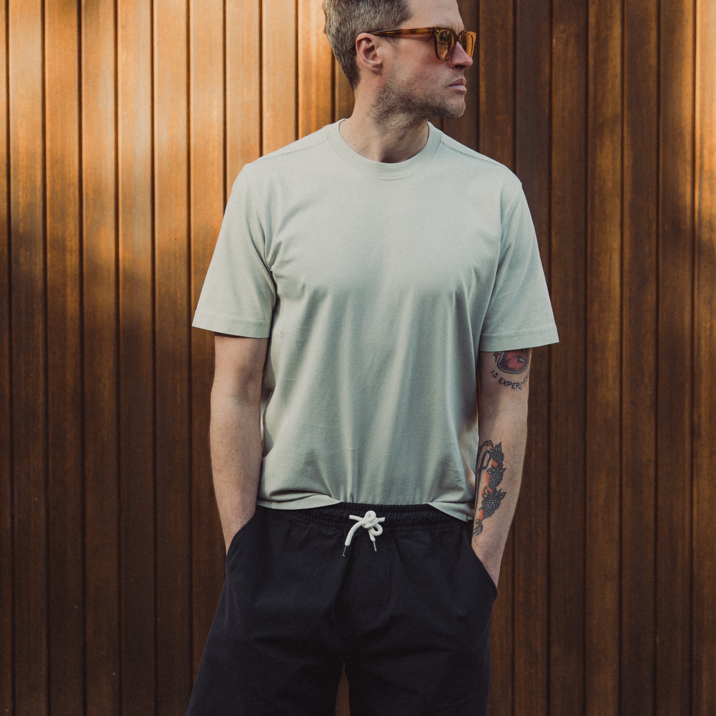 Man wearing a light green t-shirt and black shorts standing against a wooden wall.