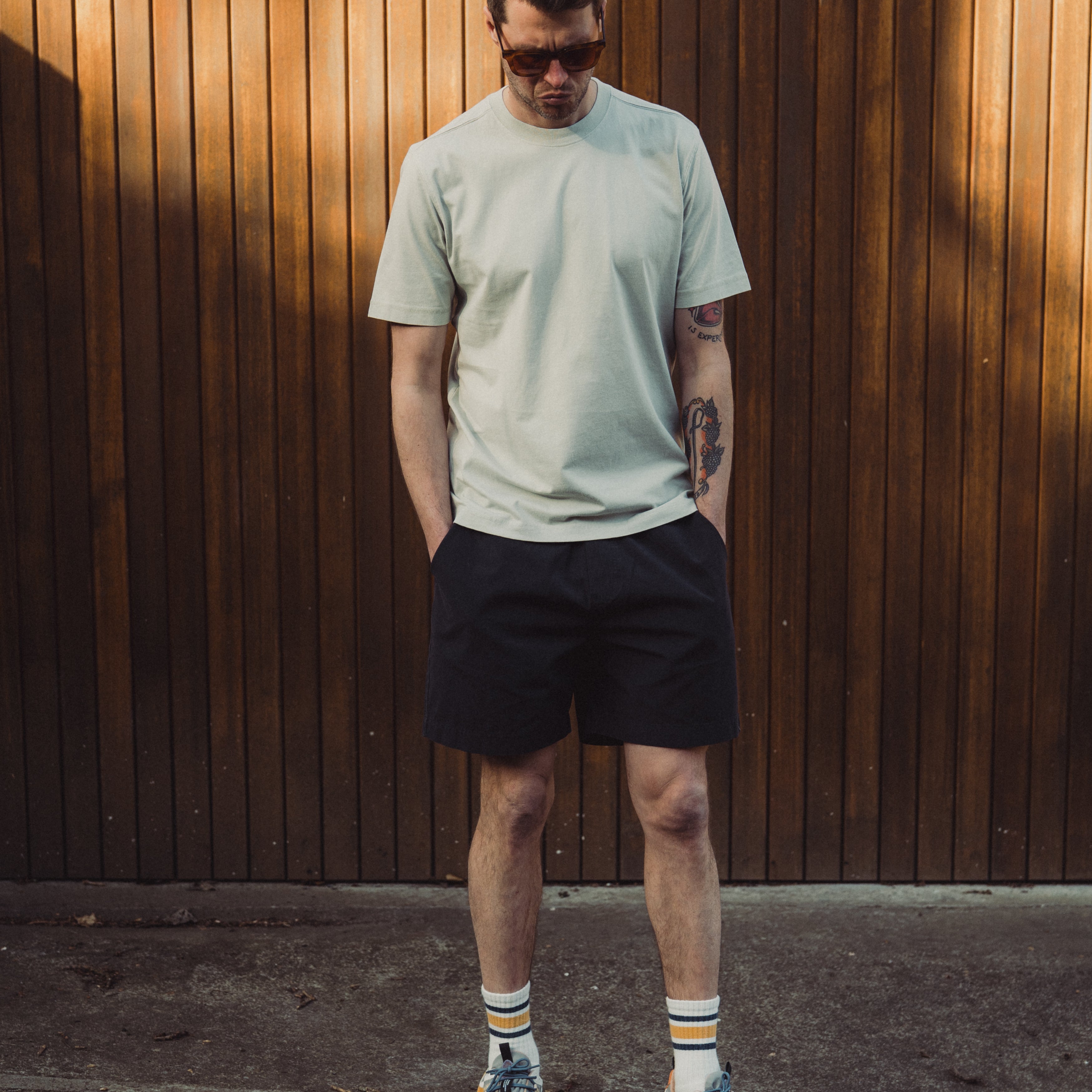 Man wearing a light green t-shirt and black shorts standing against a wooden wall.