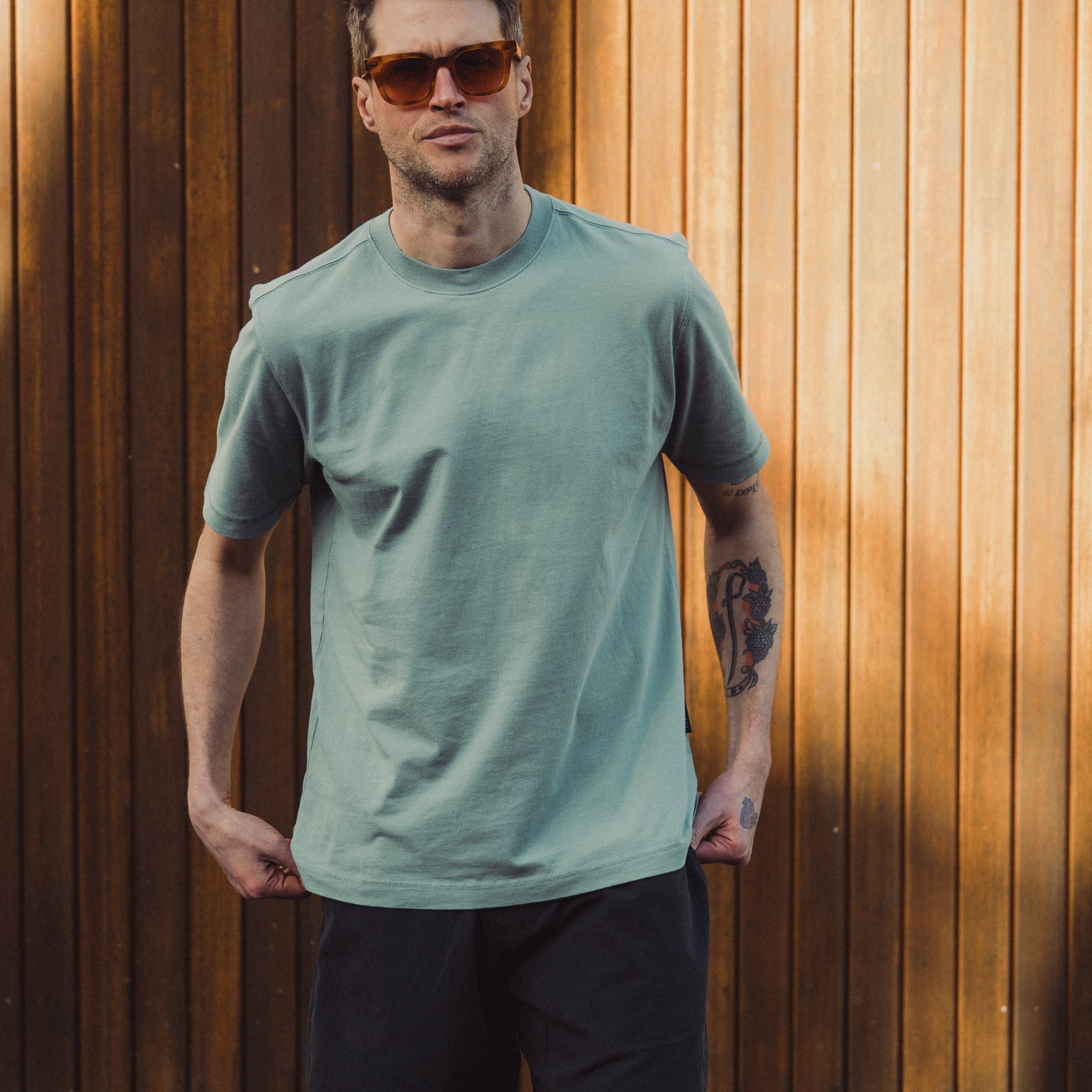 Man wearing a light green t-shirt and black shorts standing against a wooden wall.