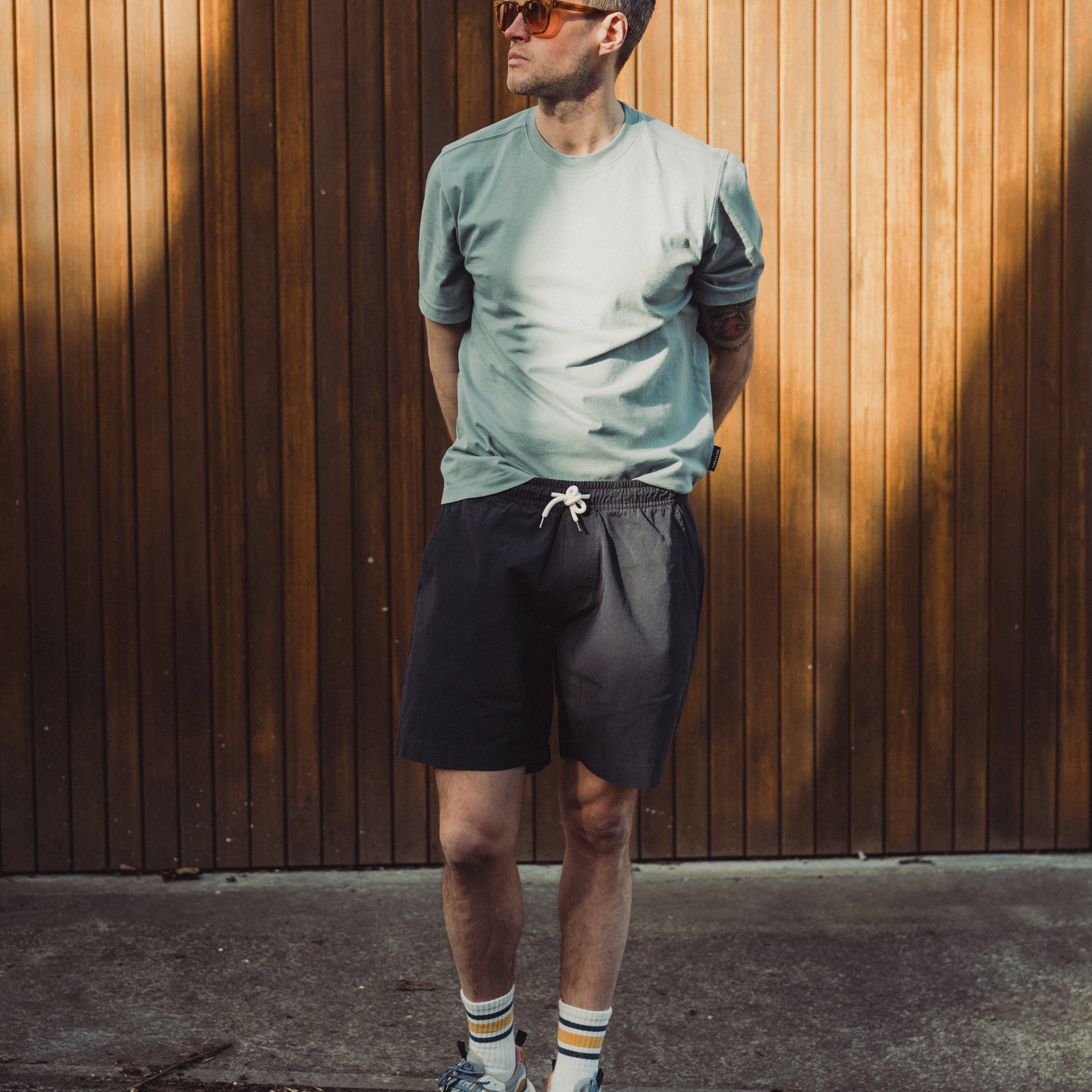 Man wearing sunglasses, a light gray t-shirt, black shorts, and colorful socks standing against a wooden wall.