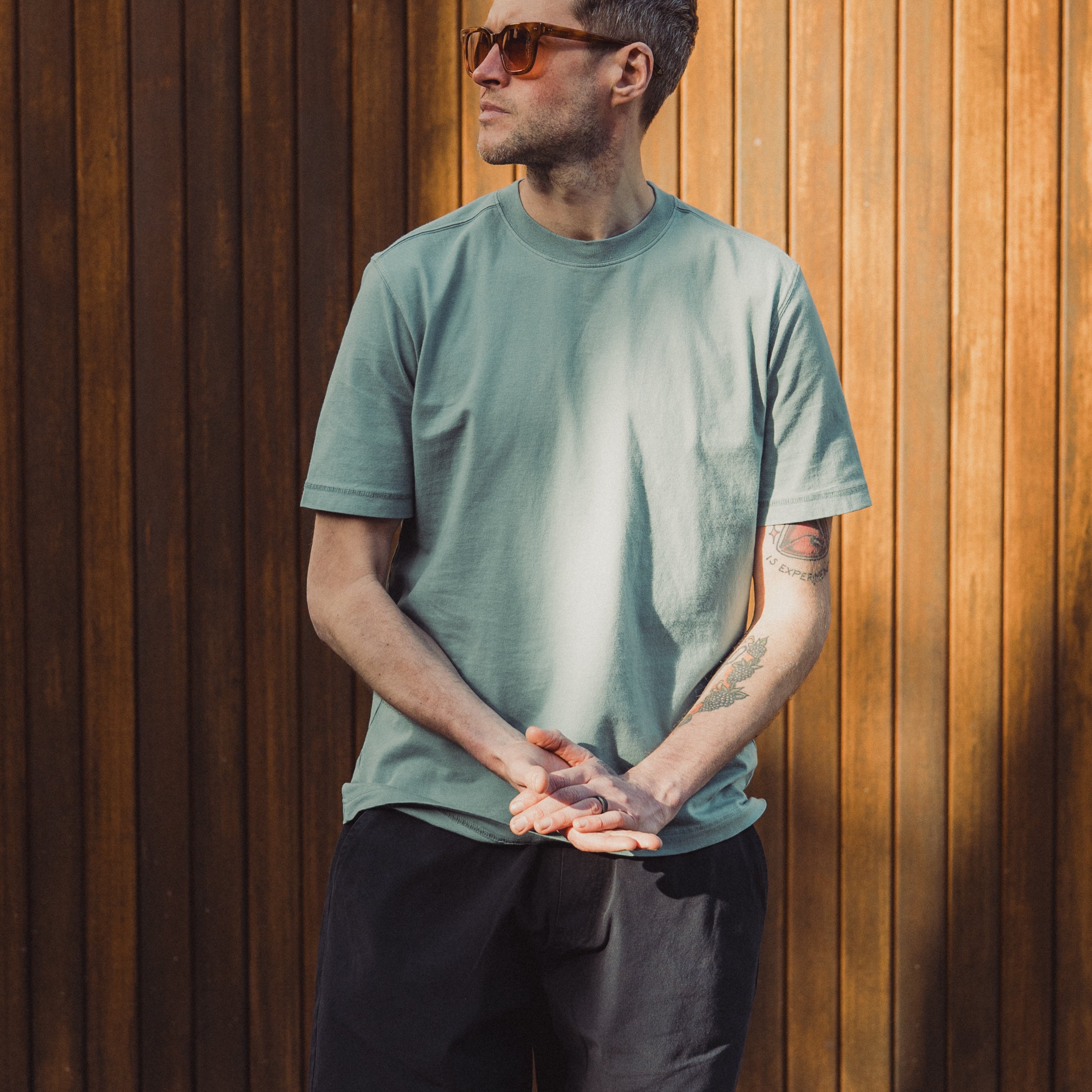 Man wearing a green t-shirt and black shorts standing against a wooden wall.