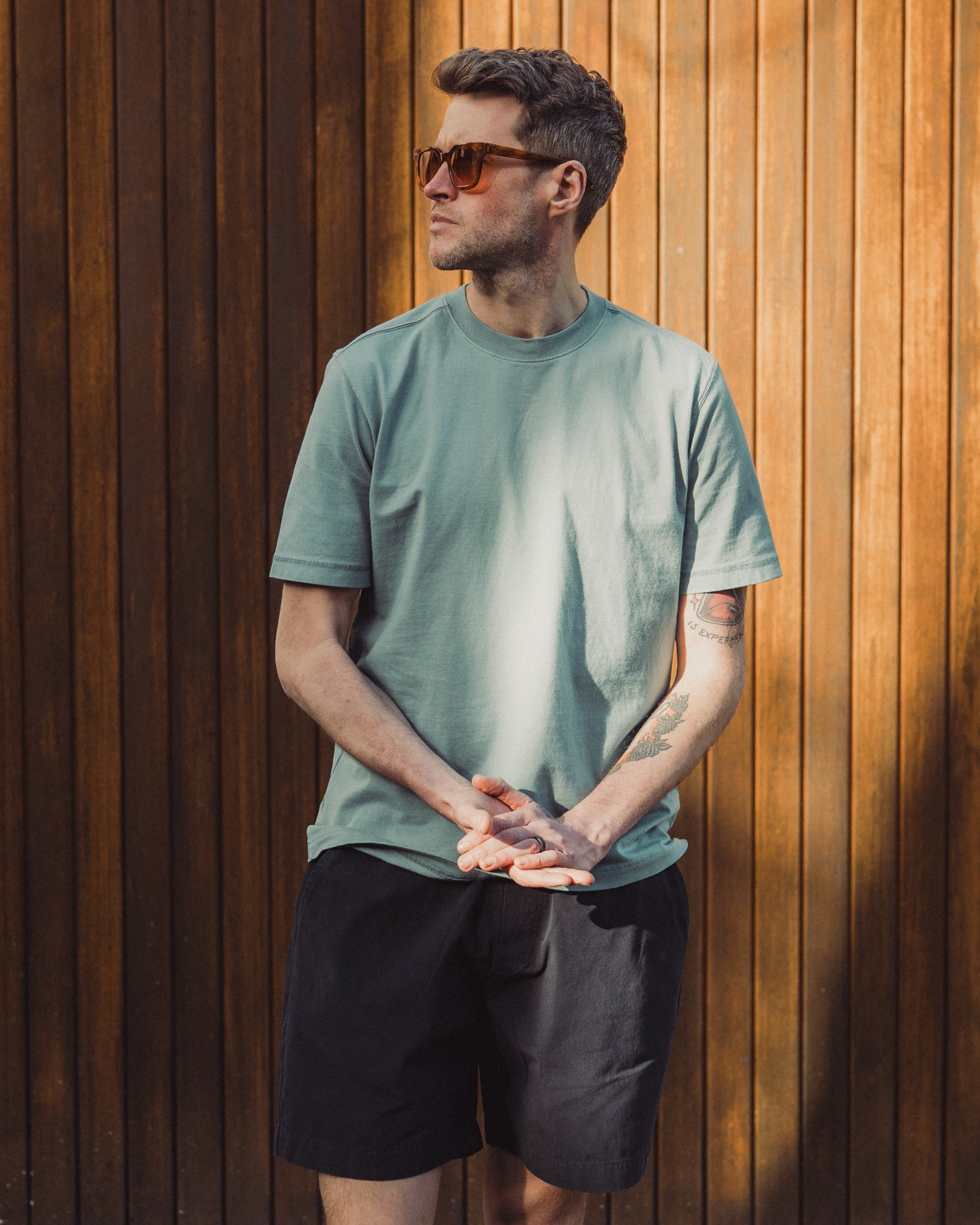 Man wearing a green t-shirt and black shorts standing against a wooden wall.