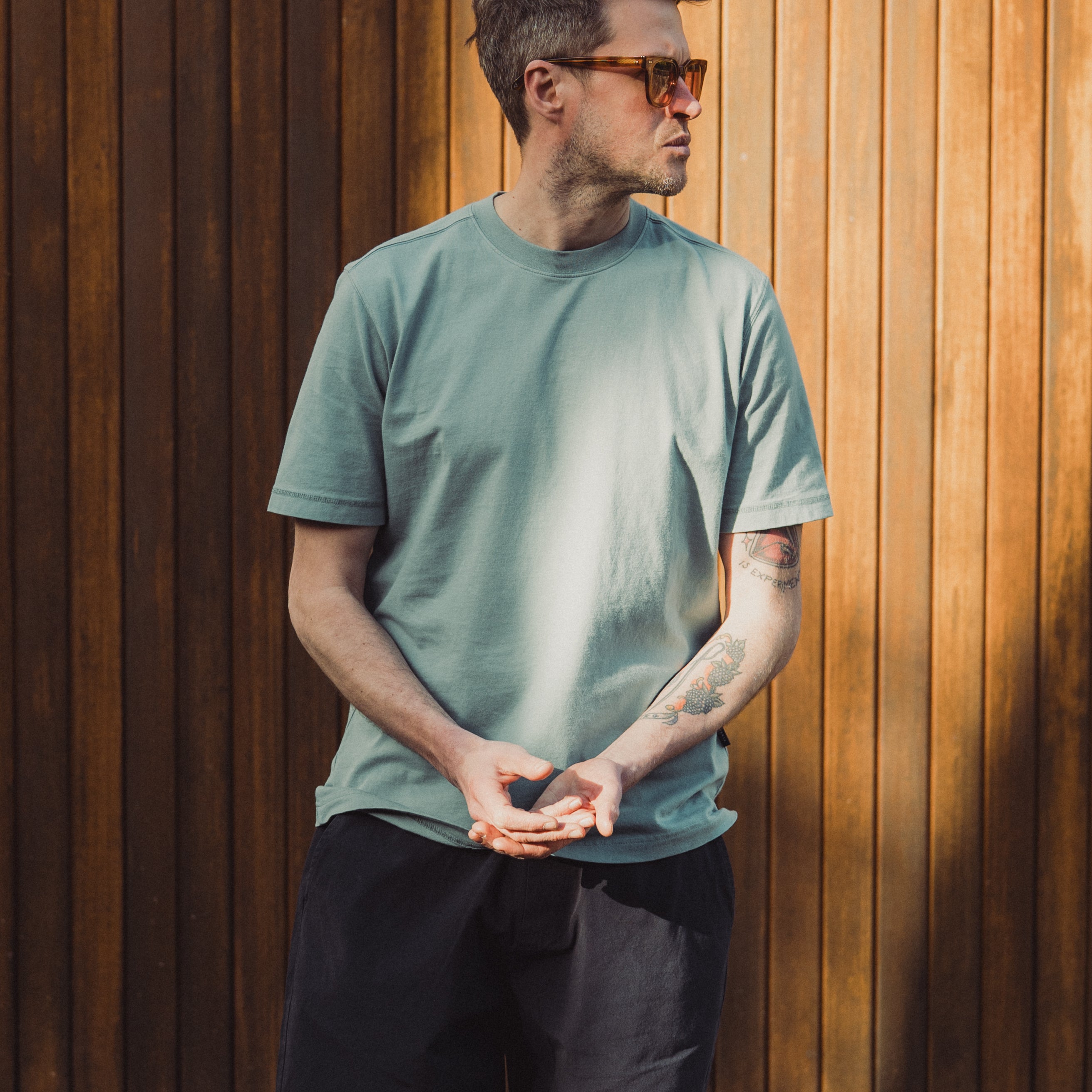 Man wearing a green t-shirt and black shorts standing against a wooden wall.