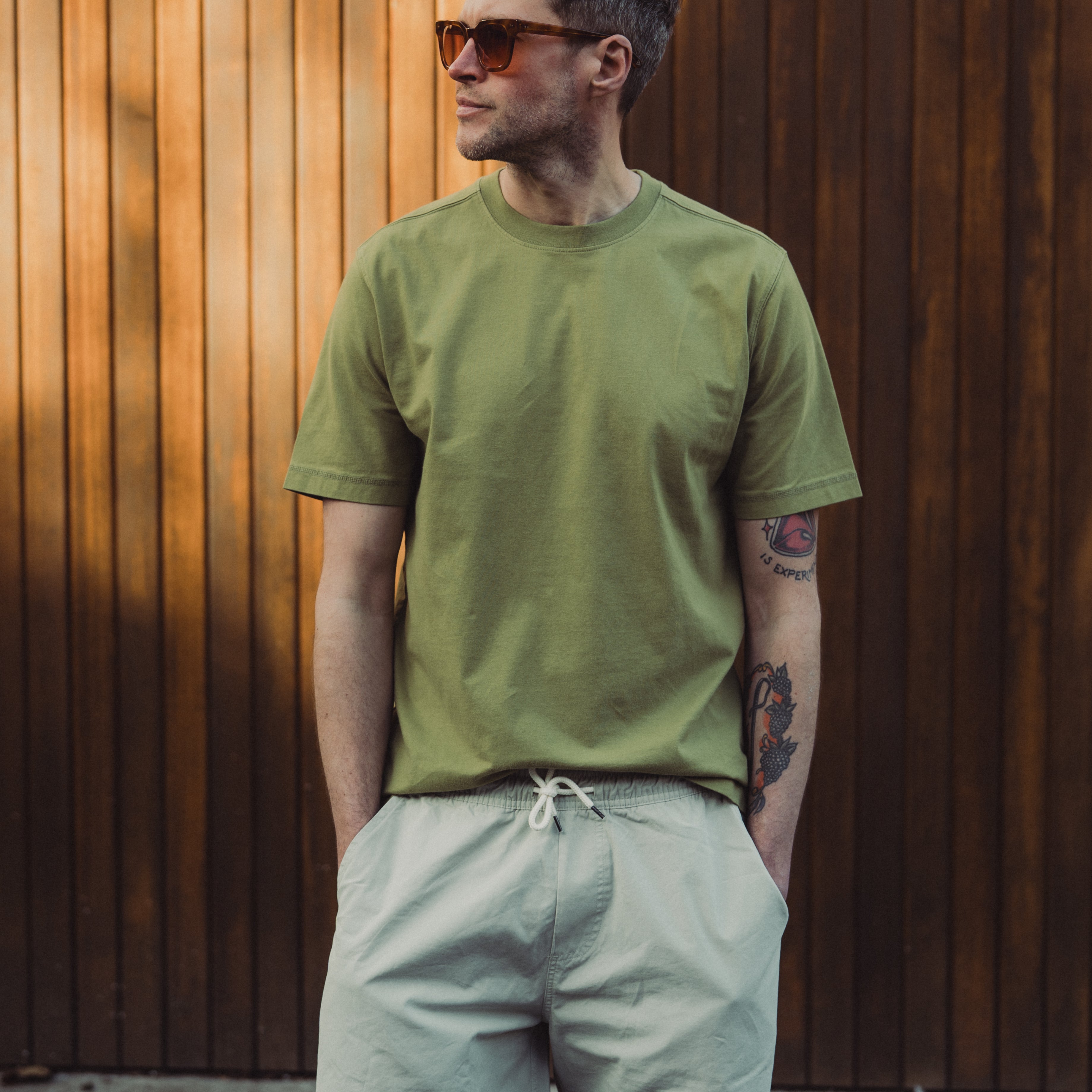 Man wearing a green t-shirt and light-colored shorts against a wooden wall.