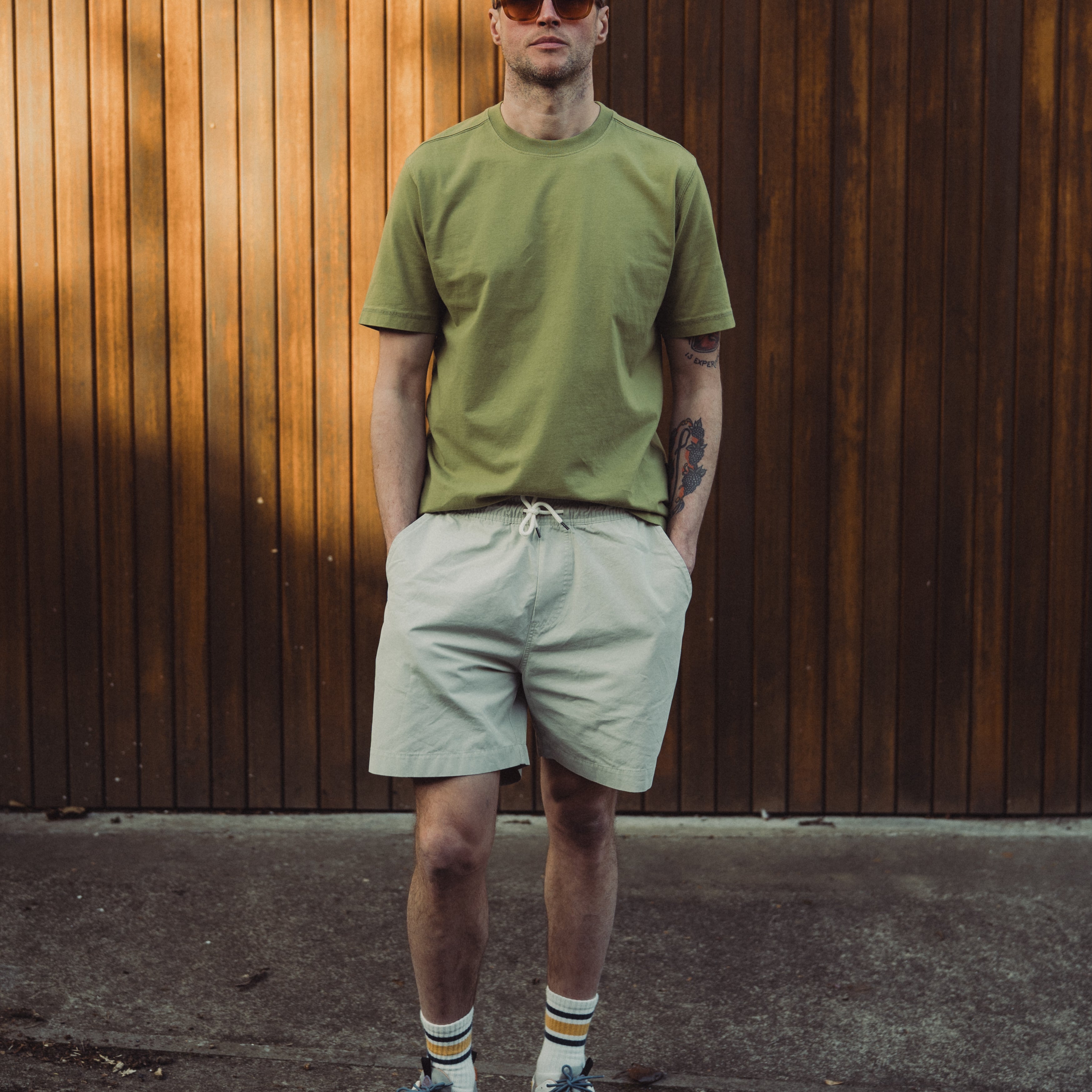Man wearing a green t-shirt and light gray shorts standing against a wooden wall.