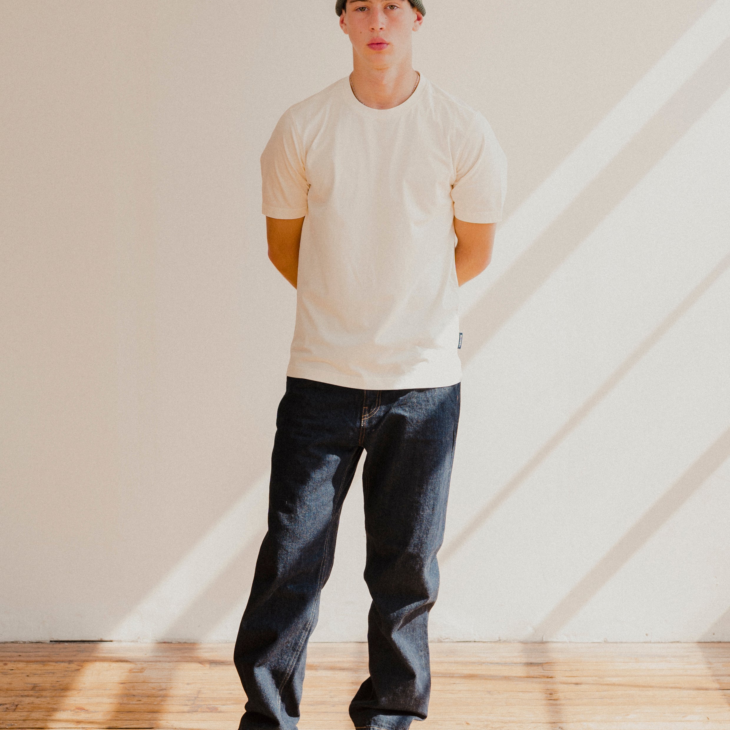 Person wearing a white t-shirt, dark jeans, and a cap standing in a room with light streaming through a window.