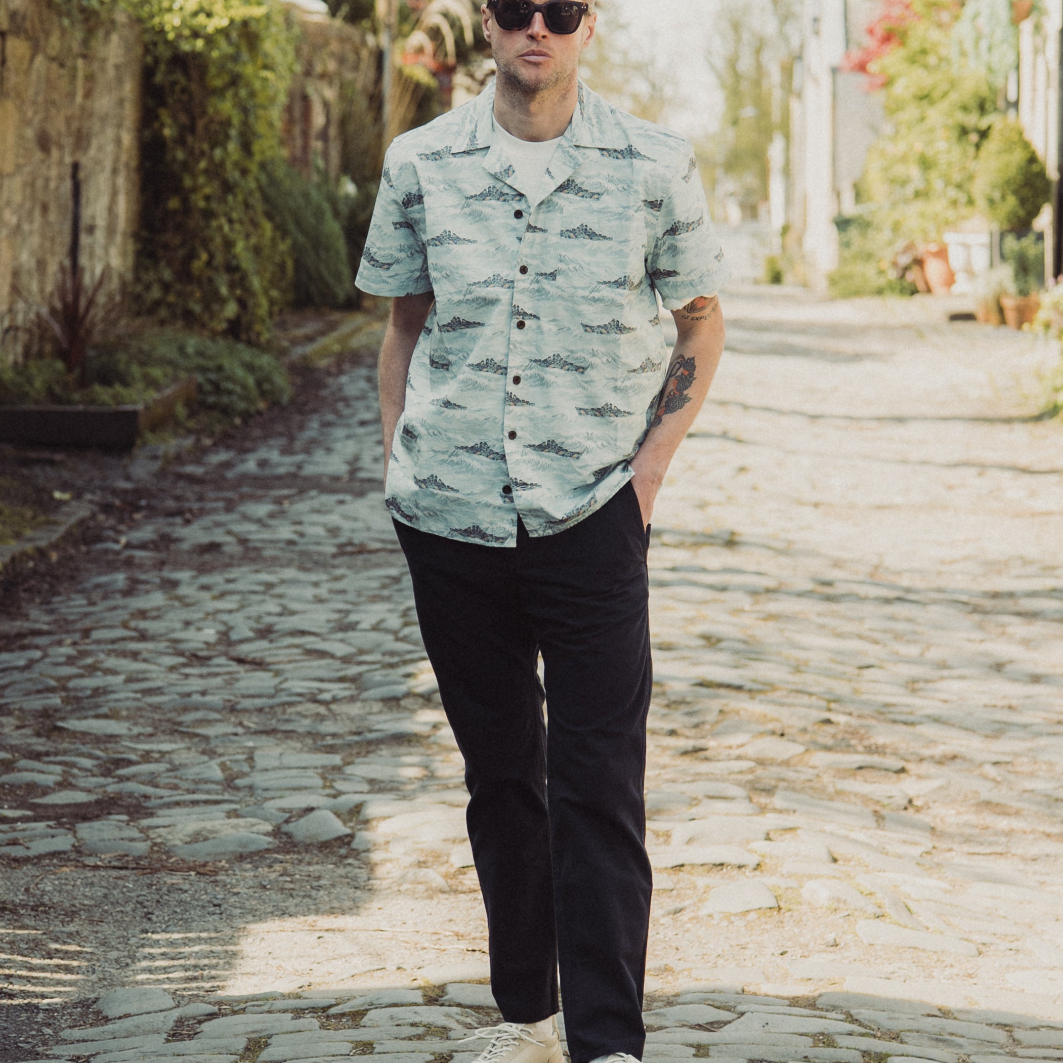 Man wearing a patterned shirt and sunglasses on a stone path with greenery.