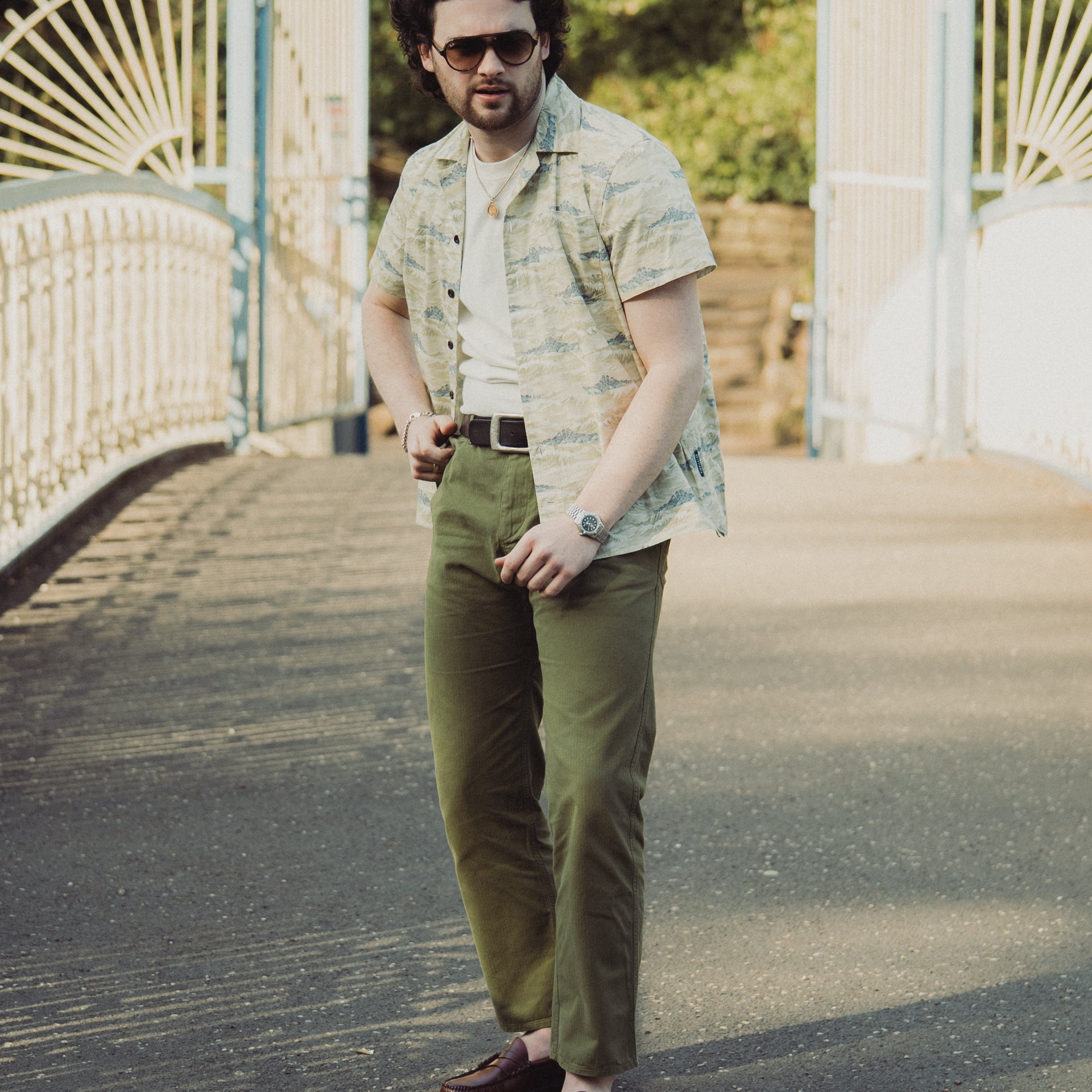 Man wearing sunglasses and a patterned shirt standing on a path with a white gate and greenery in the background.