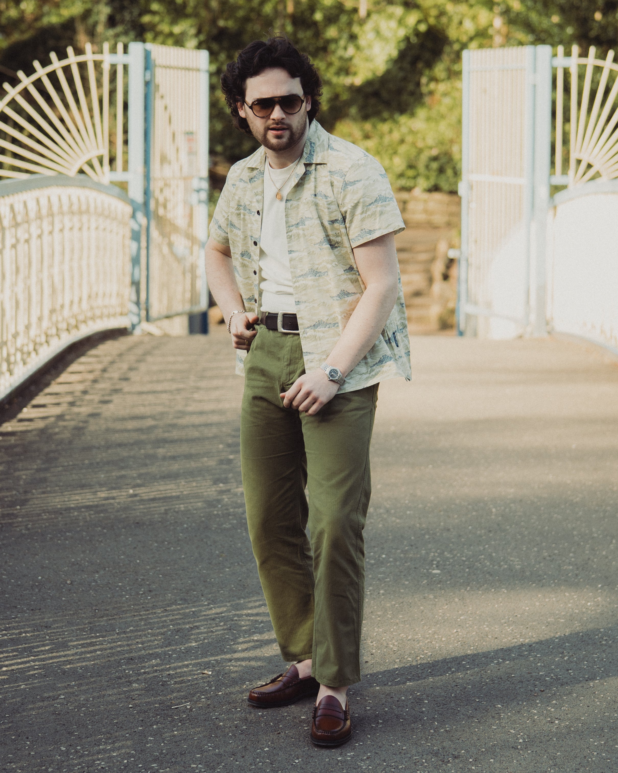 Man wearing sunglasses and a patterned shirt standing on a path with a white gate and greenery in the background.