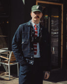 Man wearing a denim jacket, cap, and tie standing outdoors.
