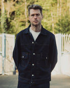 Man wearing a dark denim jacket outdoors with blurred greenery in the background