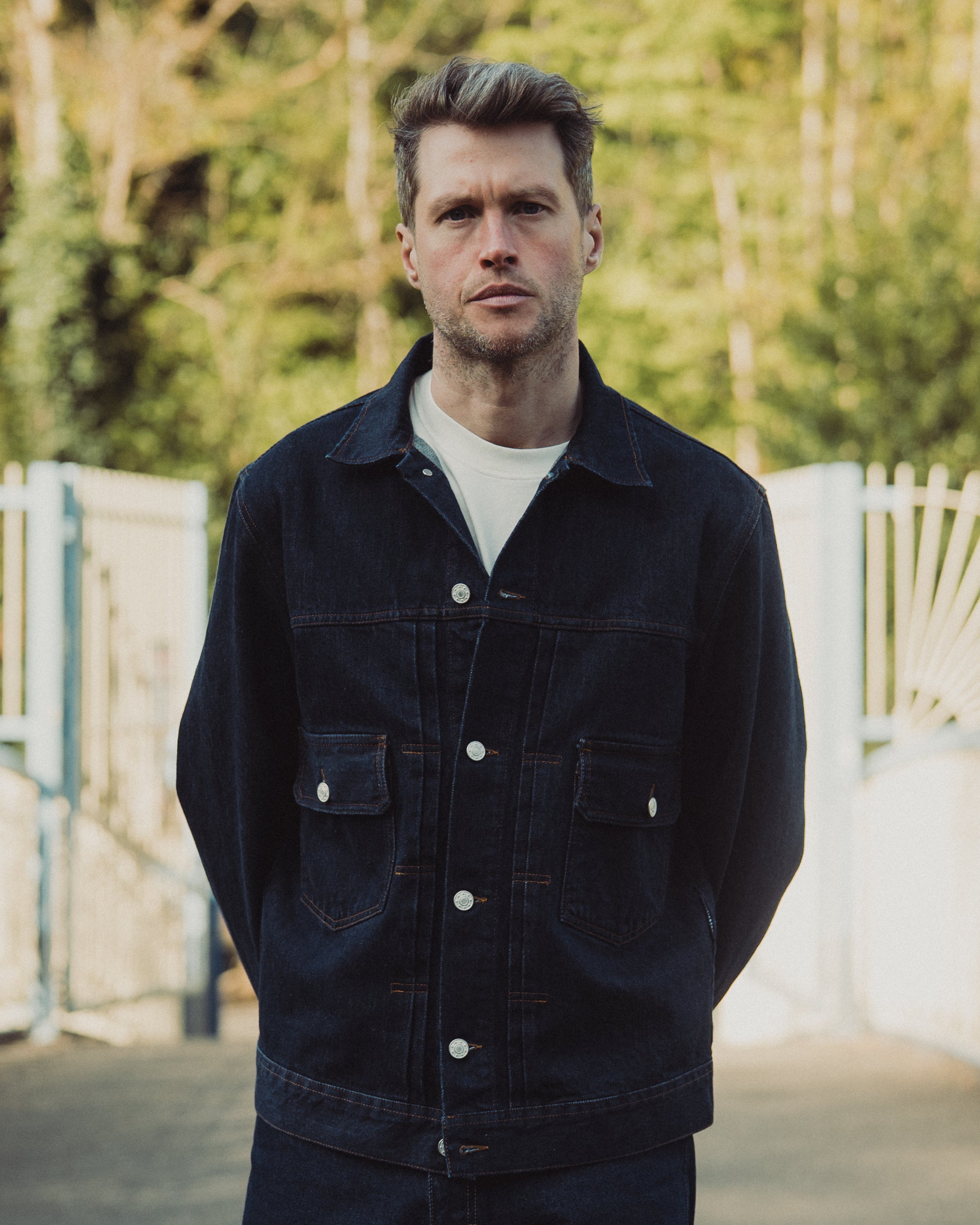 Man wearing a dark denim jacket outdoors with blurred greenery in the background