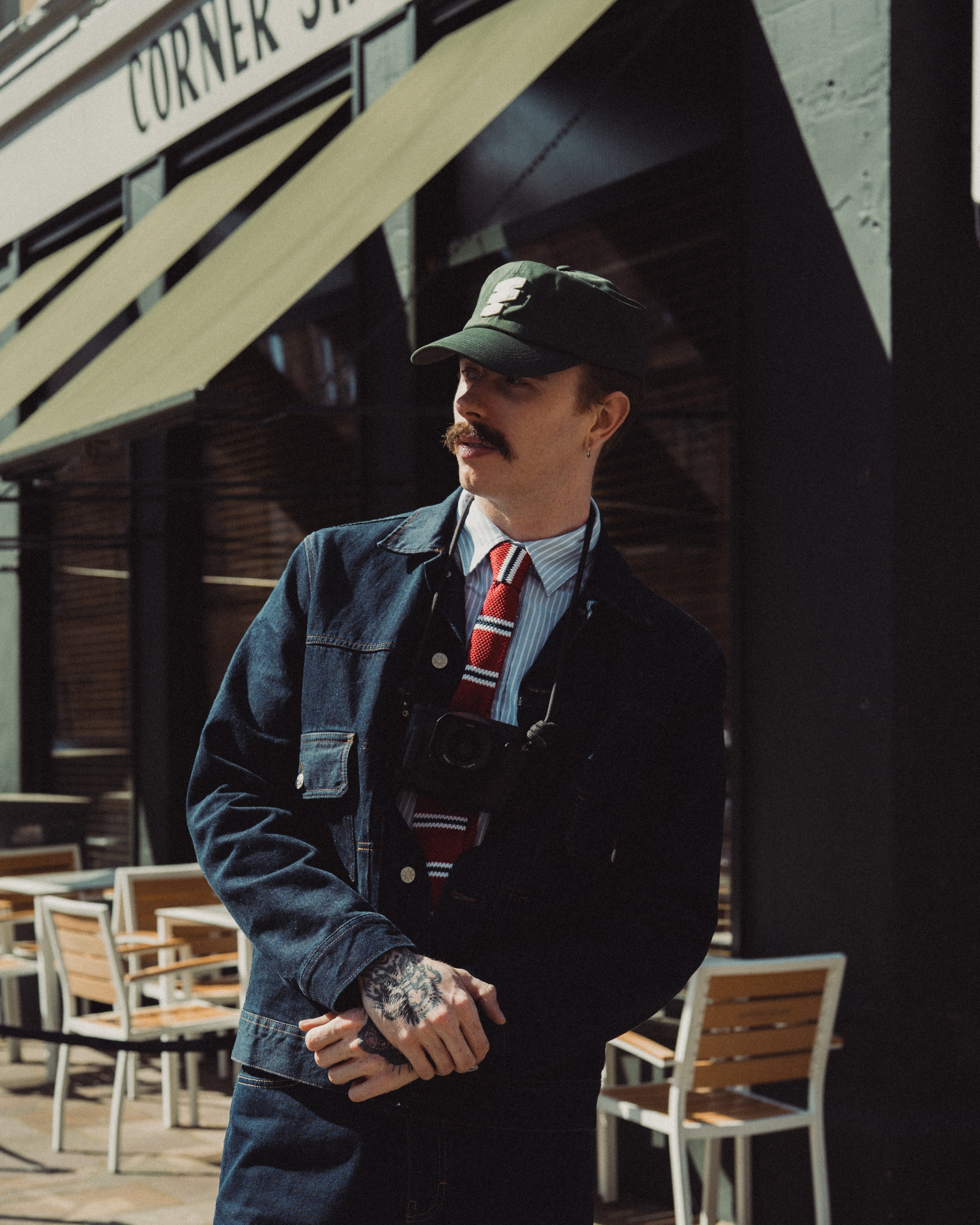 Man in a denim jacket and cap standing in front of a building with an awning.