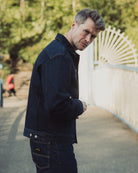 Man wearing a dark jacket and pants standing outdoors with trees and a white structure in the background.