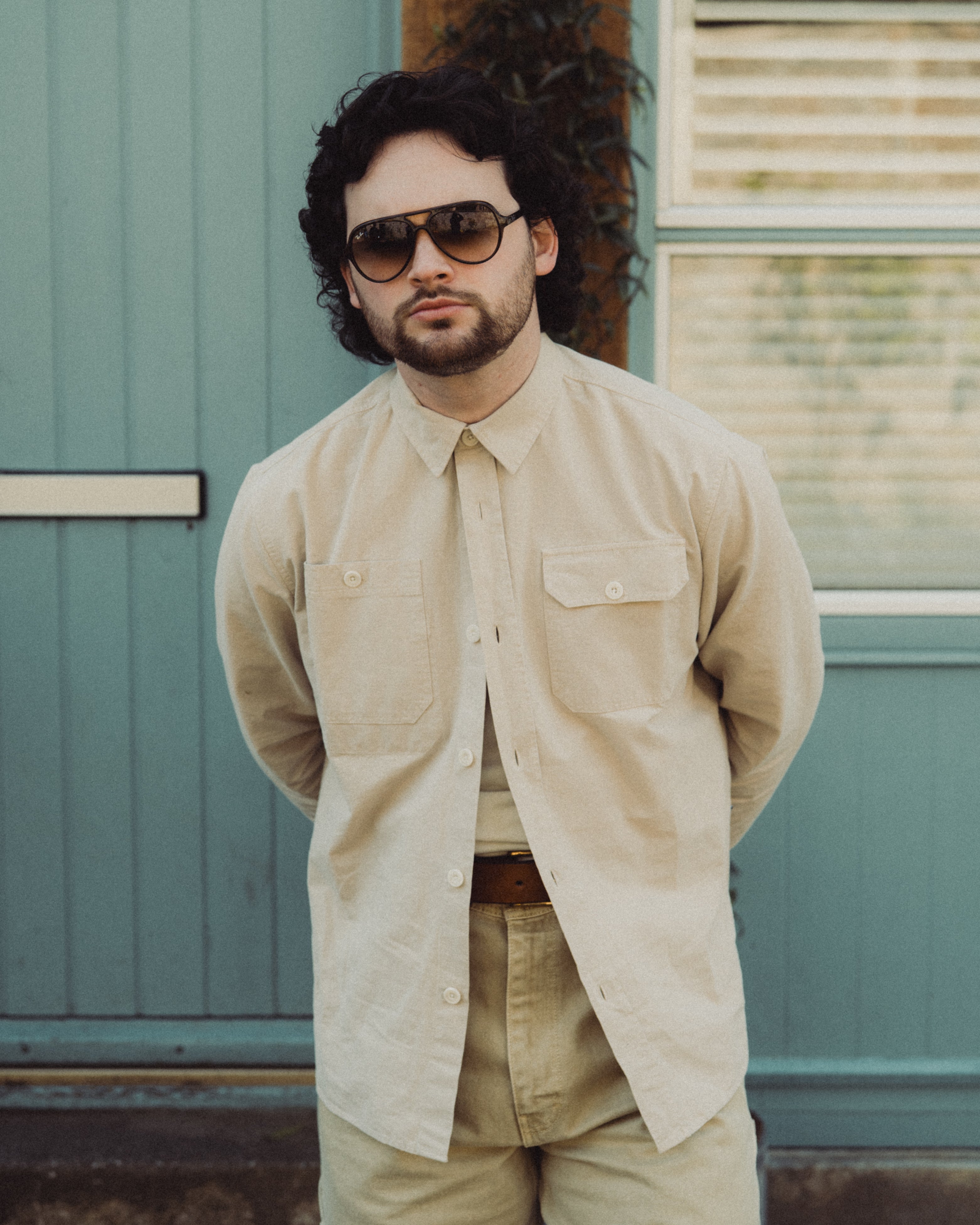 Man wearing sunglasses and a beige shirt standing in front of a teal door.