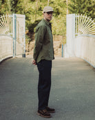 Man wearing a green jacket and cap standing on a bridge with a white gate and trees in the background.