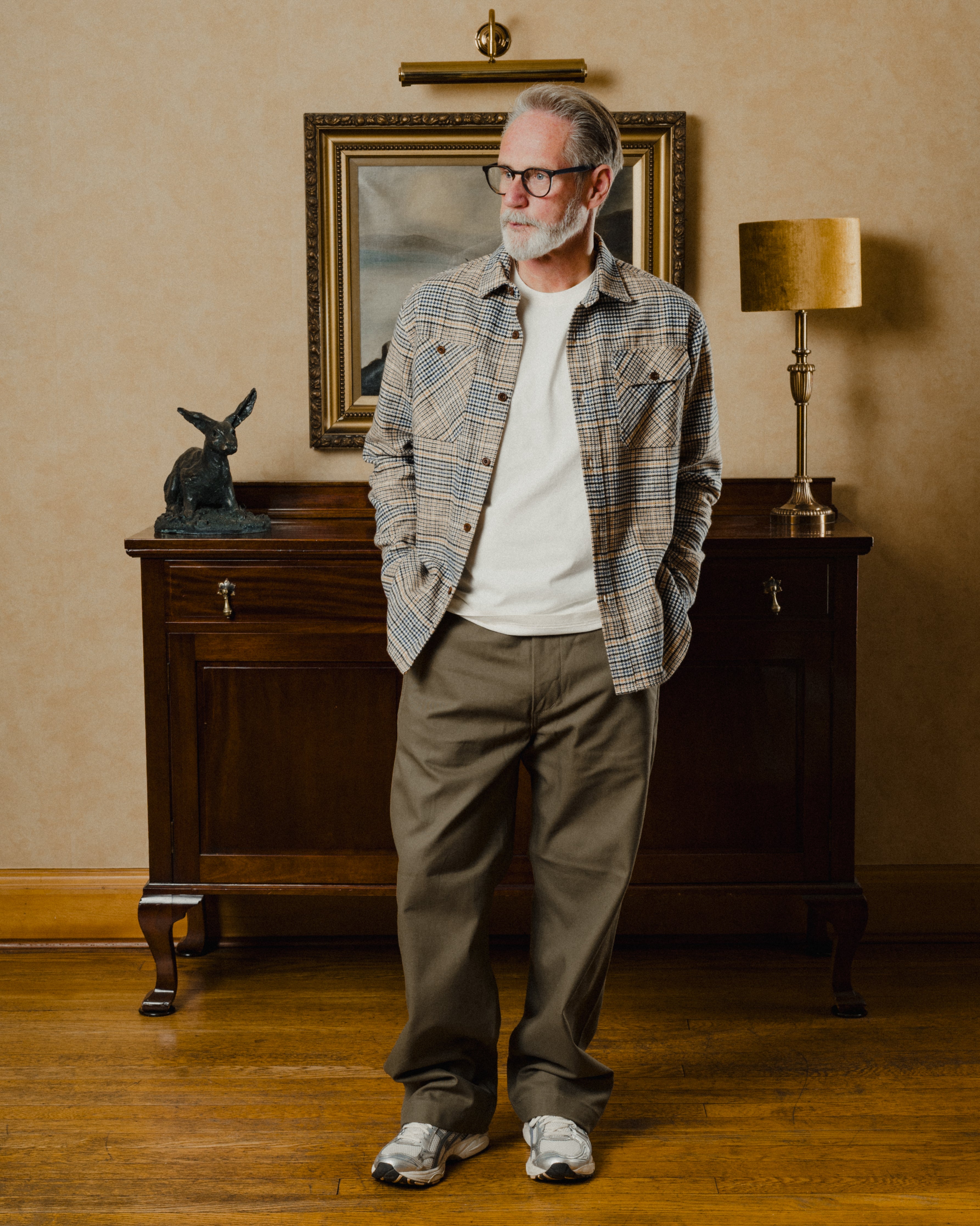 Man standing in a room with a wooden cabinet, lamp, and painting in the background.