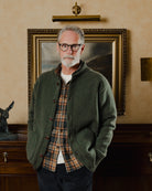 Man wearing a green cardigan over a plaid shirt in a room with a painting on the wall.