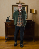 Man wearing a green cardigan over a plaid shirt in a room with a wooden cabinet and lamp.