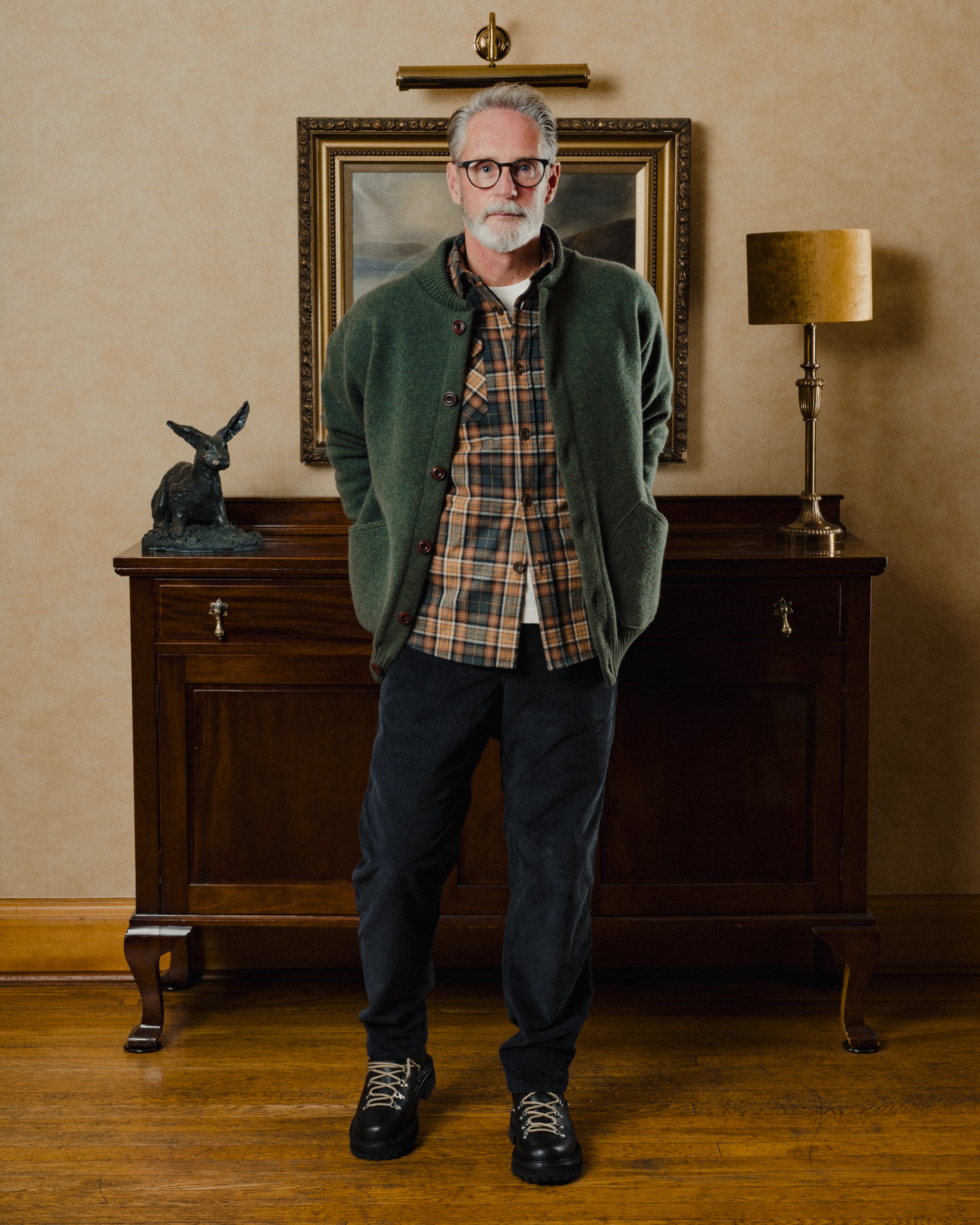 Man wearing a green cardigan over a plaid shirt in a room with a wooden cabinet and lamp.