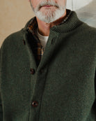 Man wearing a green coat with a plaid shirt underneath, standing against a neutral background.