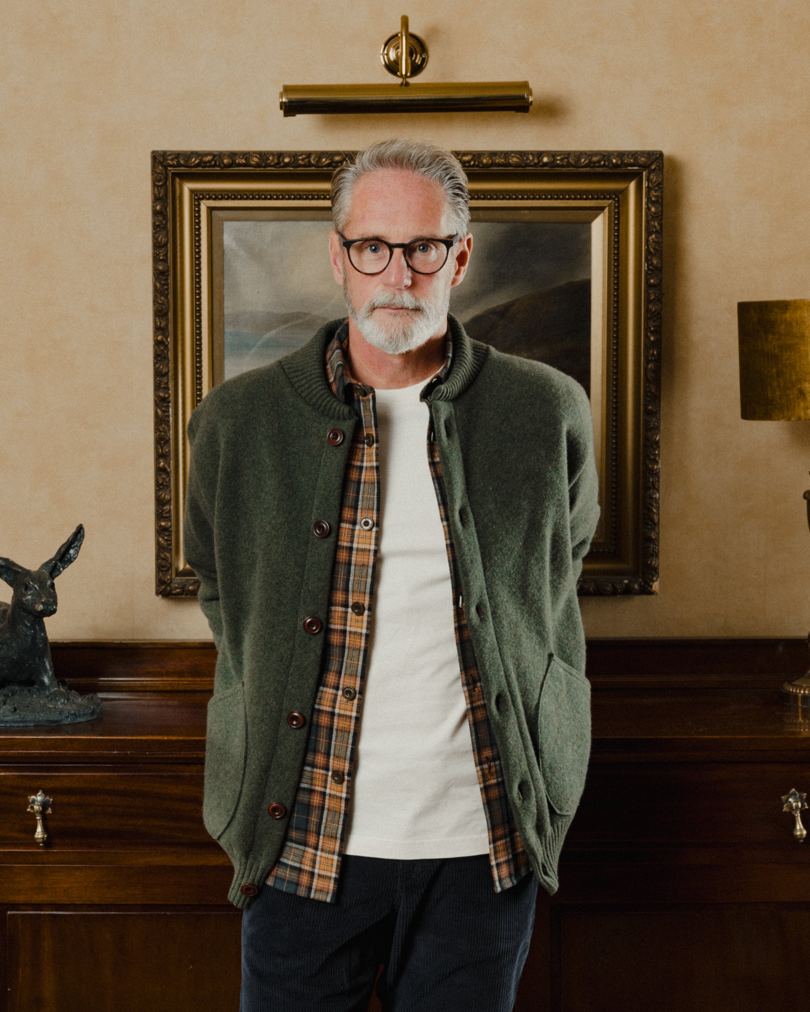 Man wearing glasses and a green jacket standing in a room with a painting on the wall.