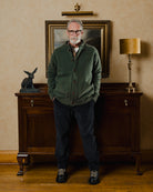 Man wearing a green sweater and dark pants standing in a room with a wooden cabinet and lamp.