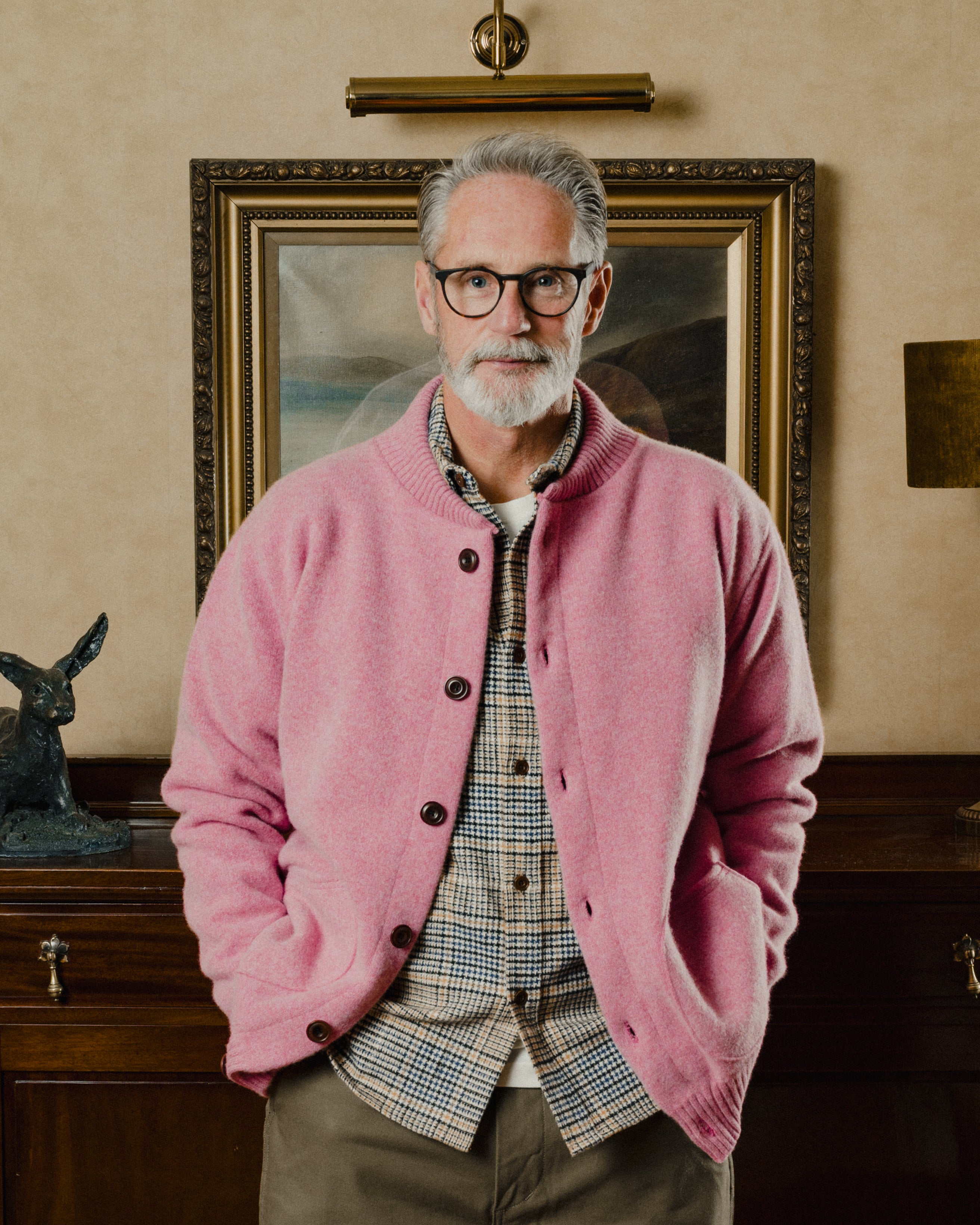 Man wearing a pink cardigan over a plaid shirt in an indoor setting with a painting in the background.