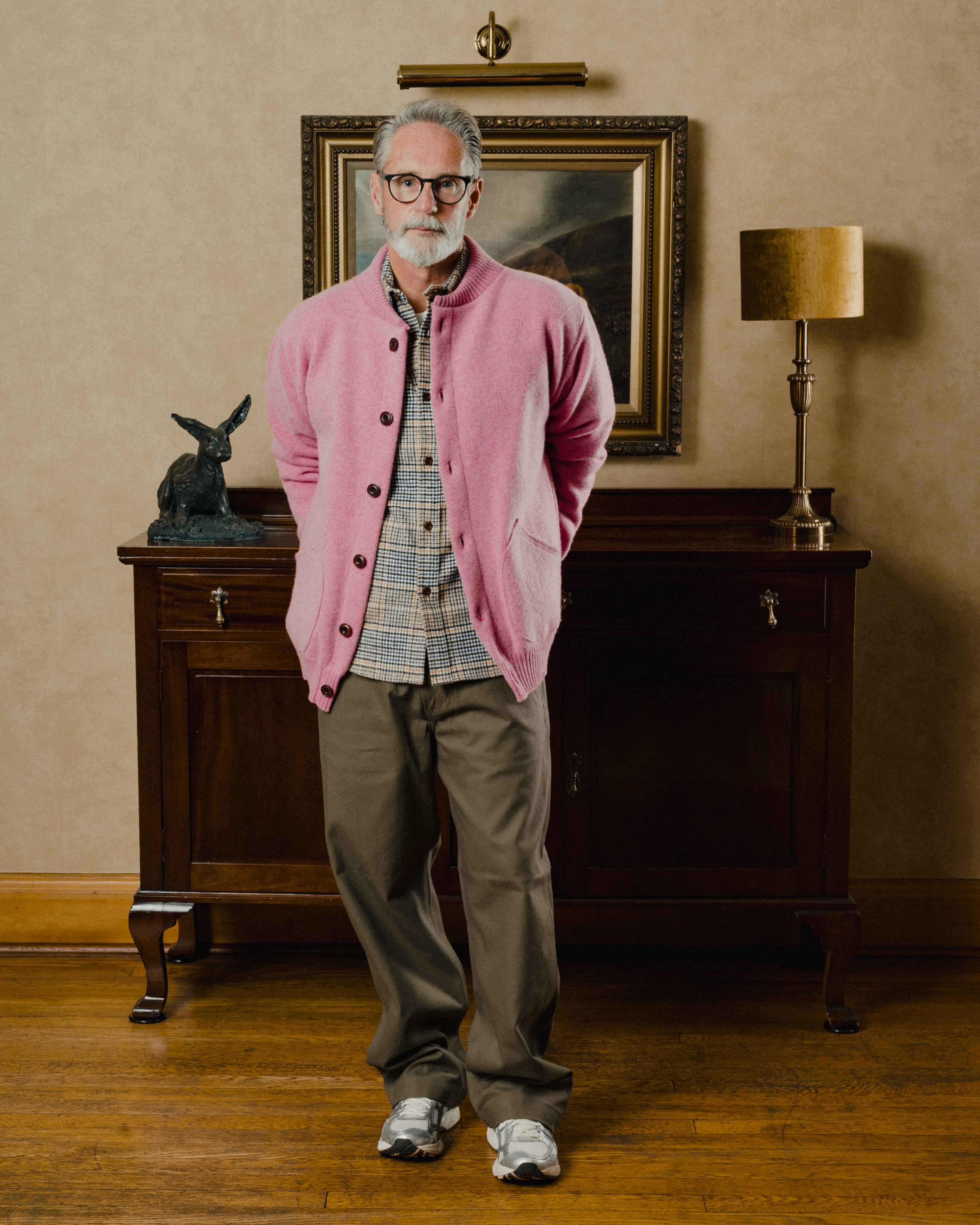 Man wearing a pink cardigan in an elegant room with a painting and lamp.