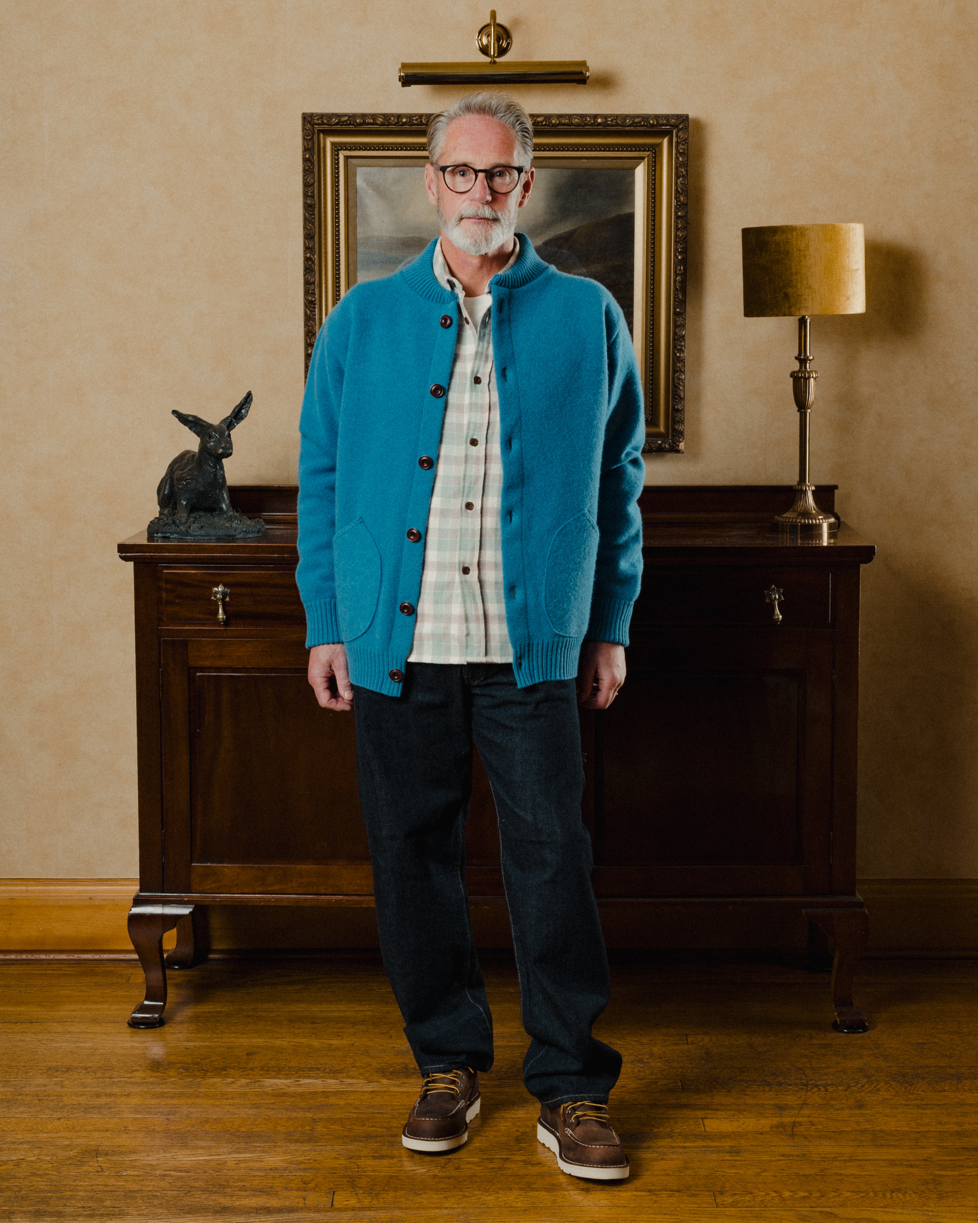Man wearing a blue cardigan in an indoor setting with a lamp and painting in the background.