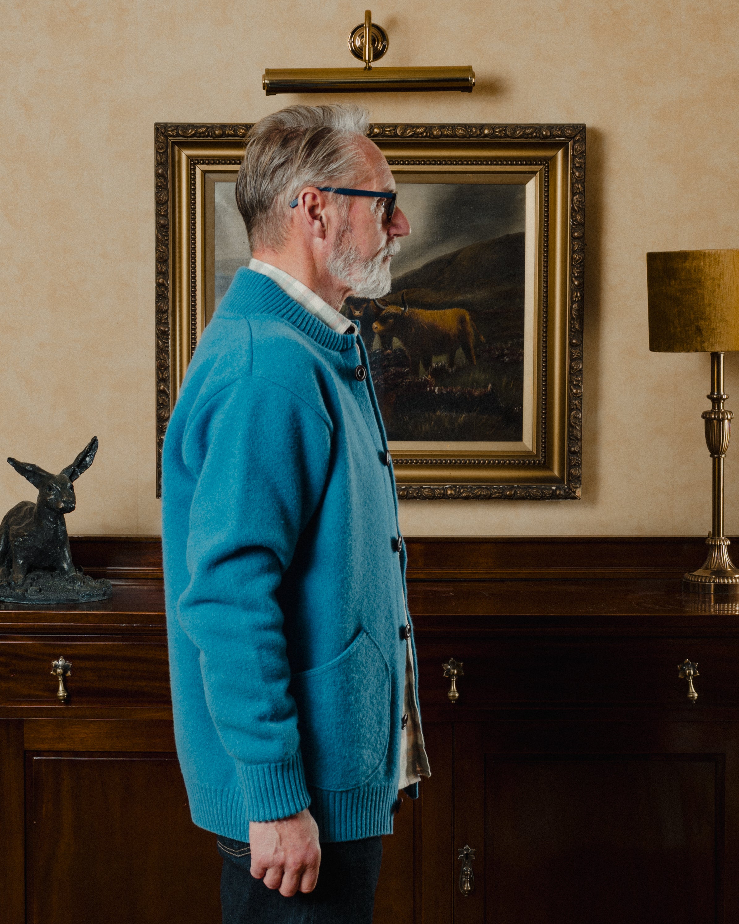 Man in a blue cardigan standing in a room with a painting of a cow on the wall.