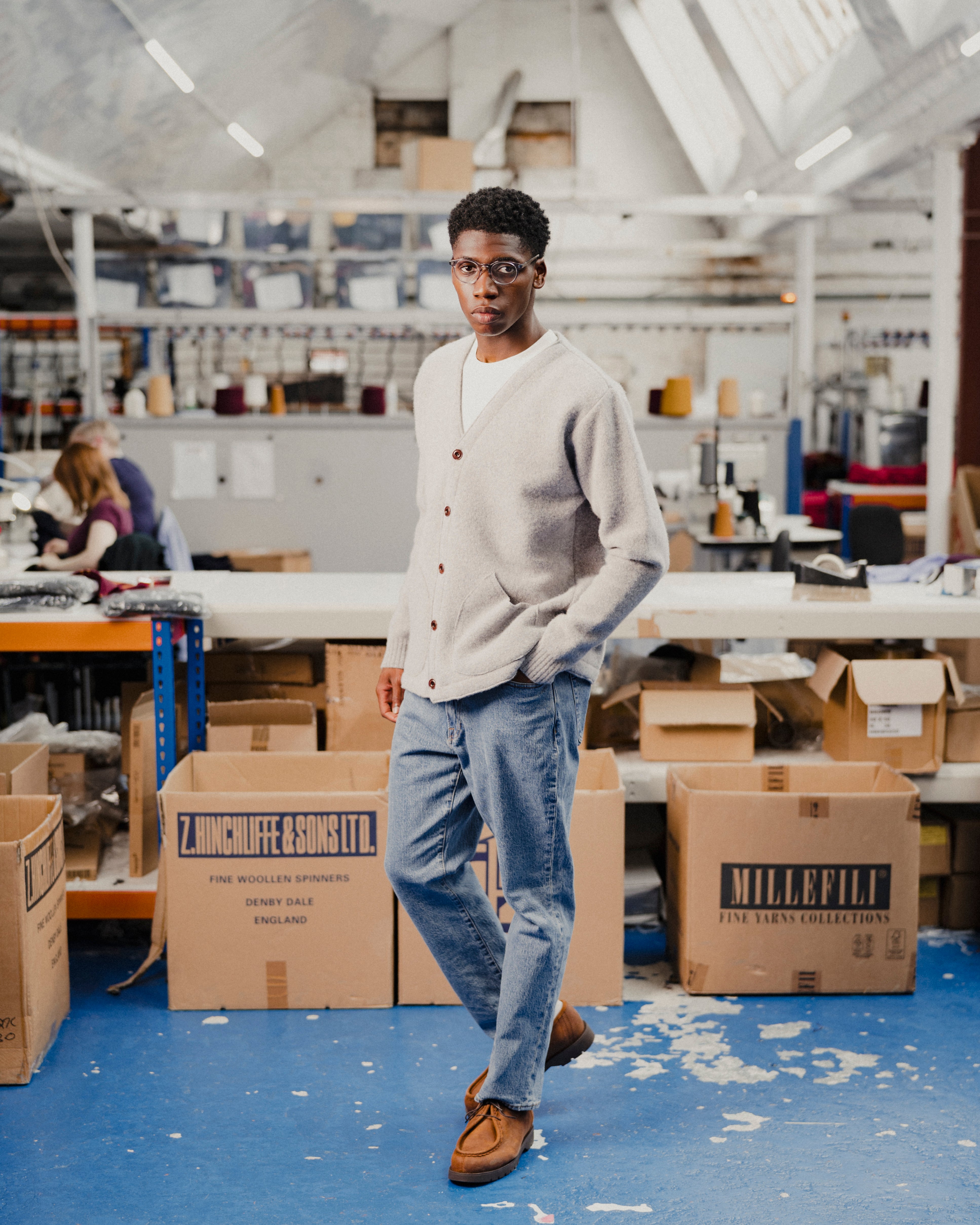 Man wearing a light grey cardigan in a fashion warehouse, stood around boxes