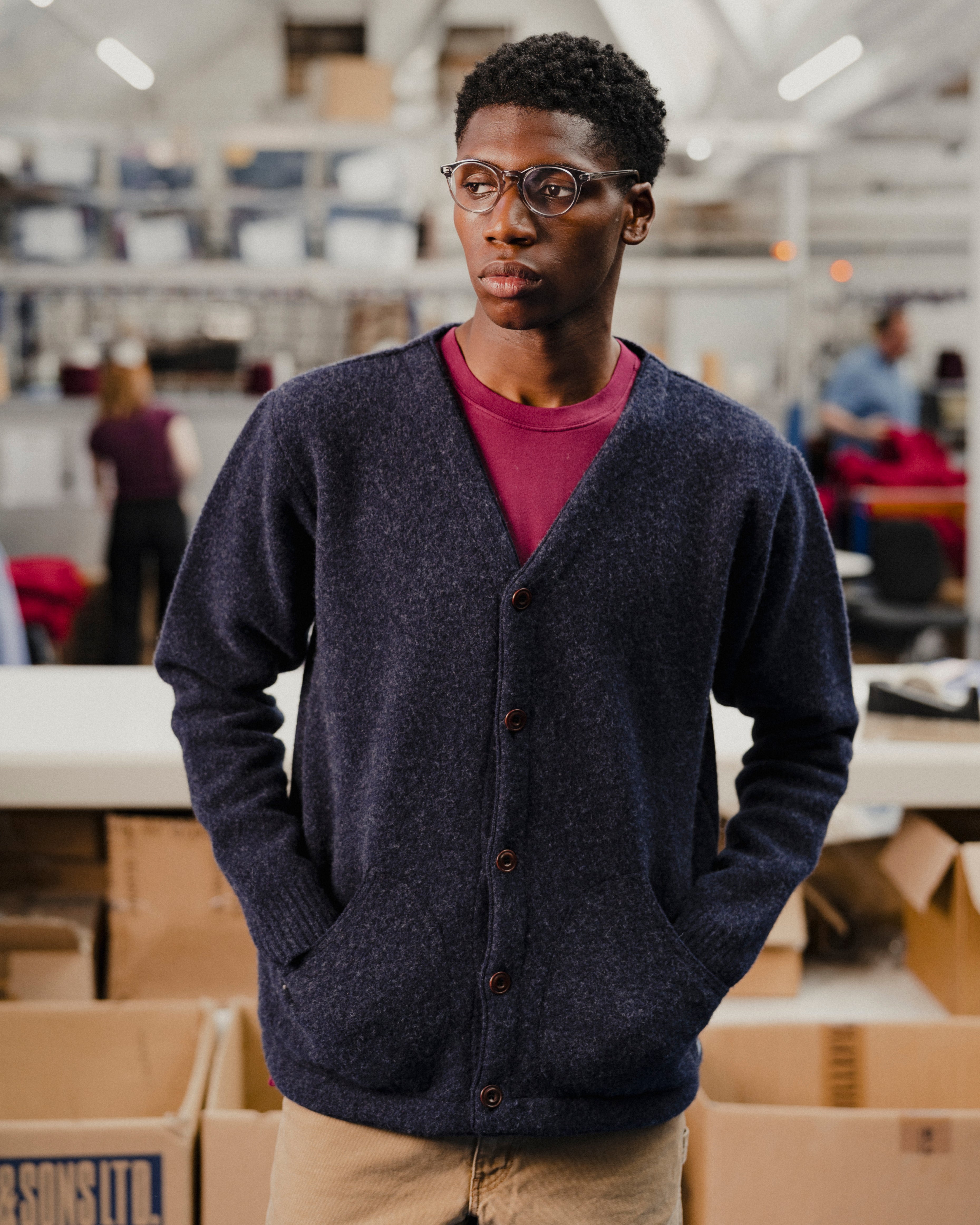 Person wearing a dark blue cardigan over a red shirt in an indoor setting with boxes and people in the background.