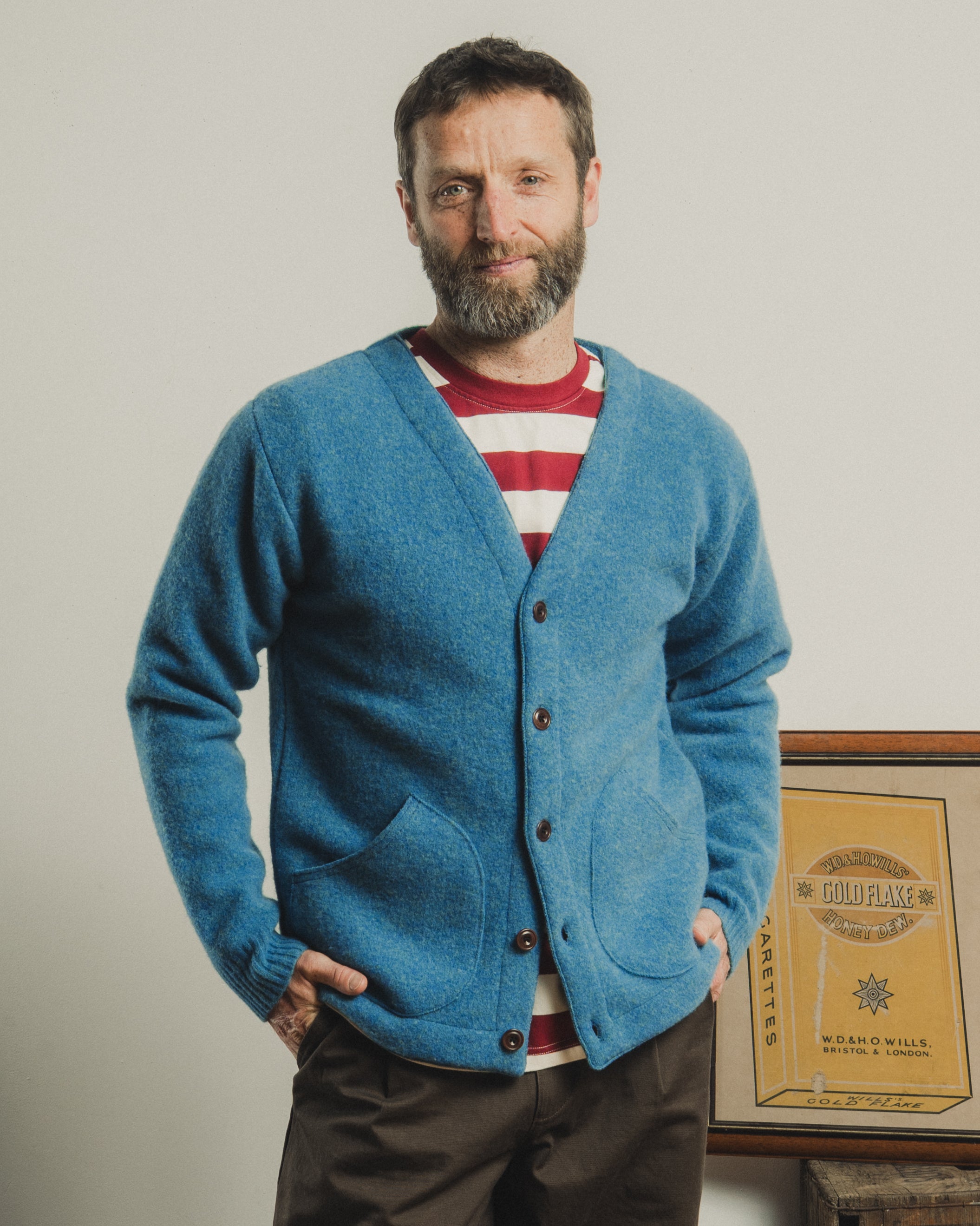Man wearing a blue cardigan over a red and white striped shirt, standing against a plain background.