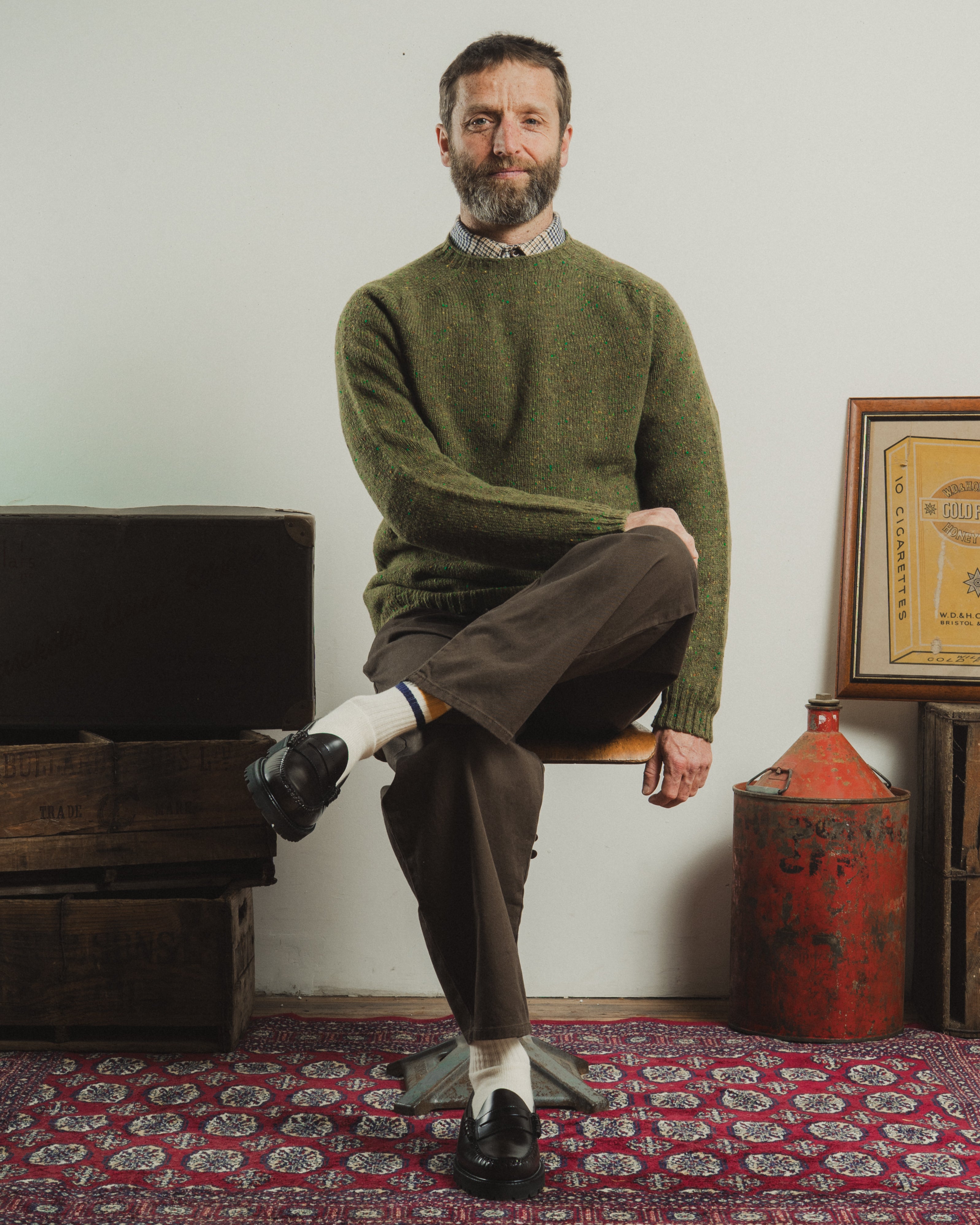 Man sitting on a chair wearing a green sweater and brown pants in a room with rustic decor.