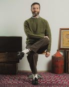 Man sitting on a chair wearing a green sweater and brown pants in a room with rustic decor.
