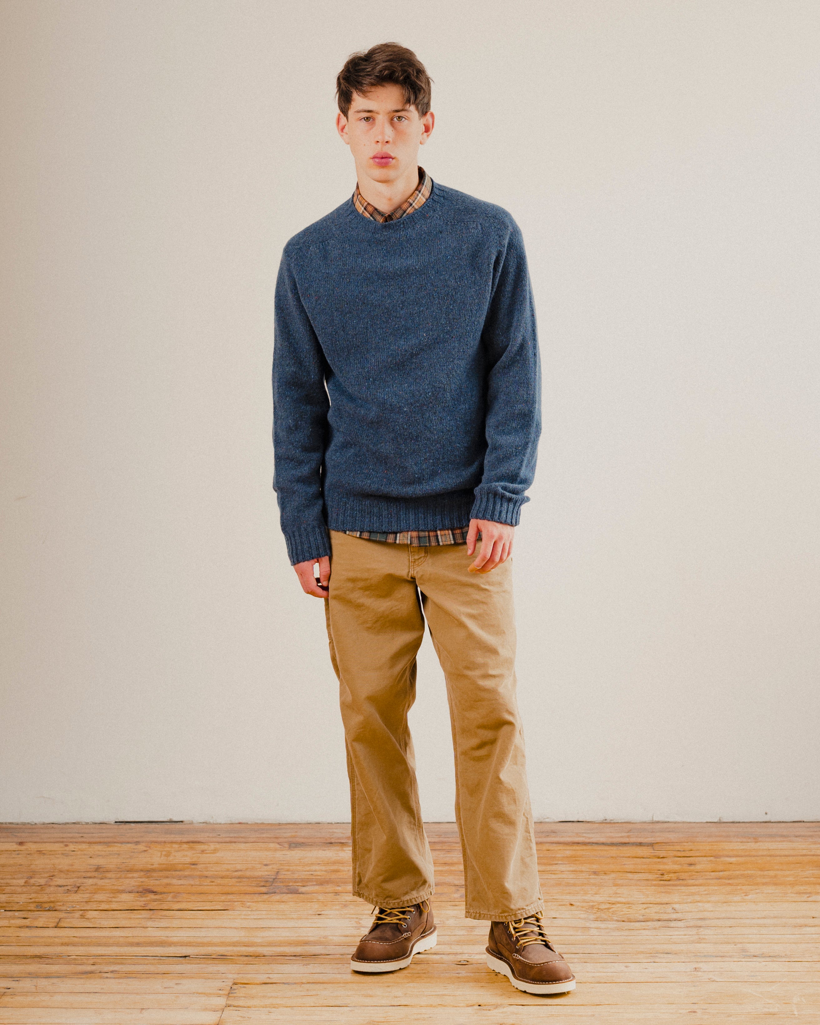 Man wearing a blue wool sweater knit crewneck with a check shirt underneath on a plain background with a wooden floor