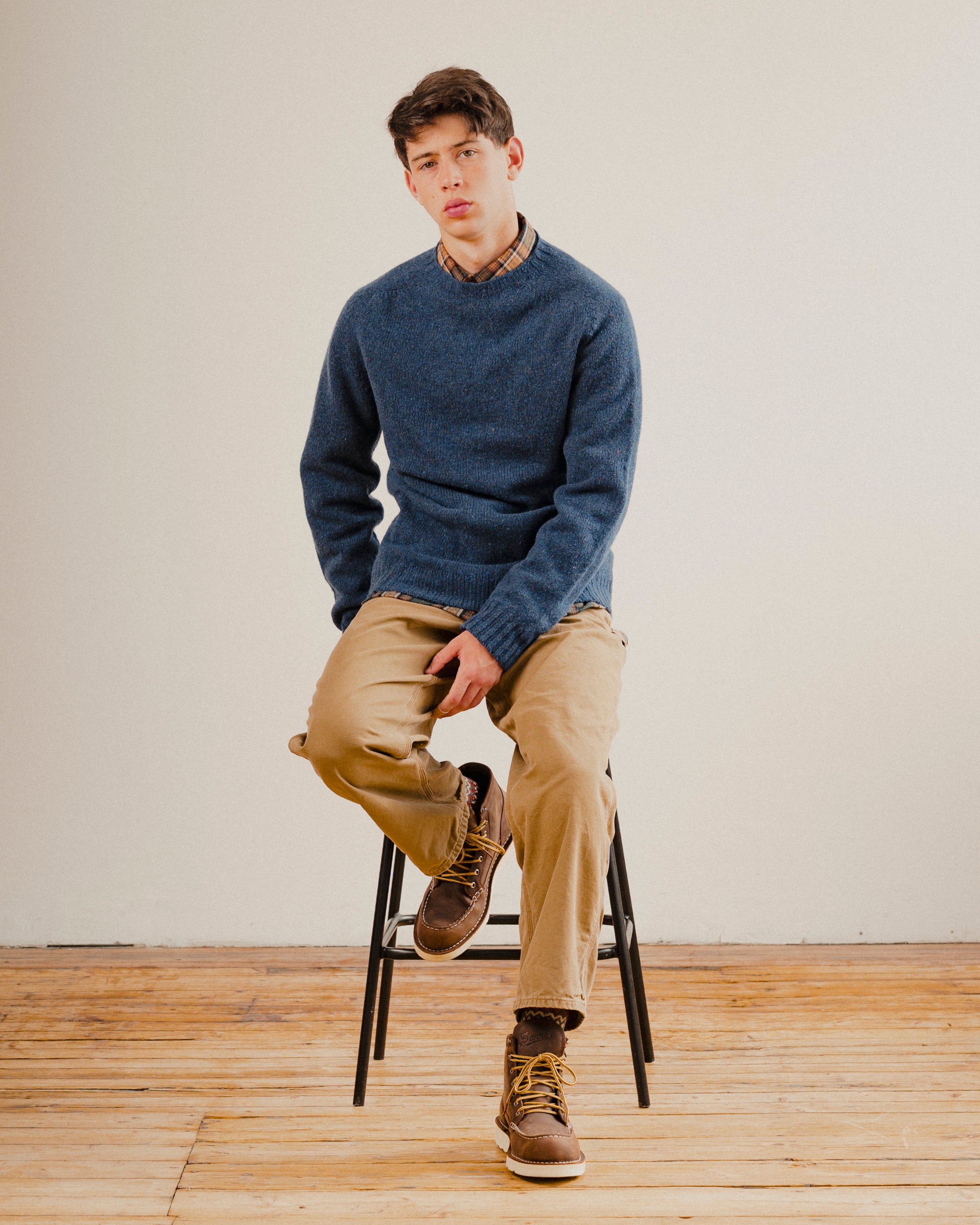 Man sat on a stool wearing a navy blue wool sweater knit crewneck with a check shirt underneath