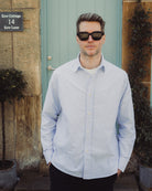Man wearing sunglasses and a light blue striped shirt standing in front of a building with a door and plants.
